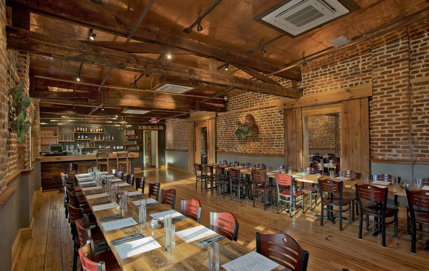 A restaurant interior with brick walls, wooden beams, and wooden floors. There are multiple tables set with menus, glasses, and utensils. The bar area is in the back with bottles on shelves.