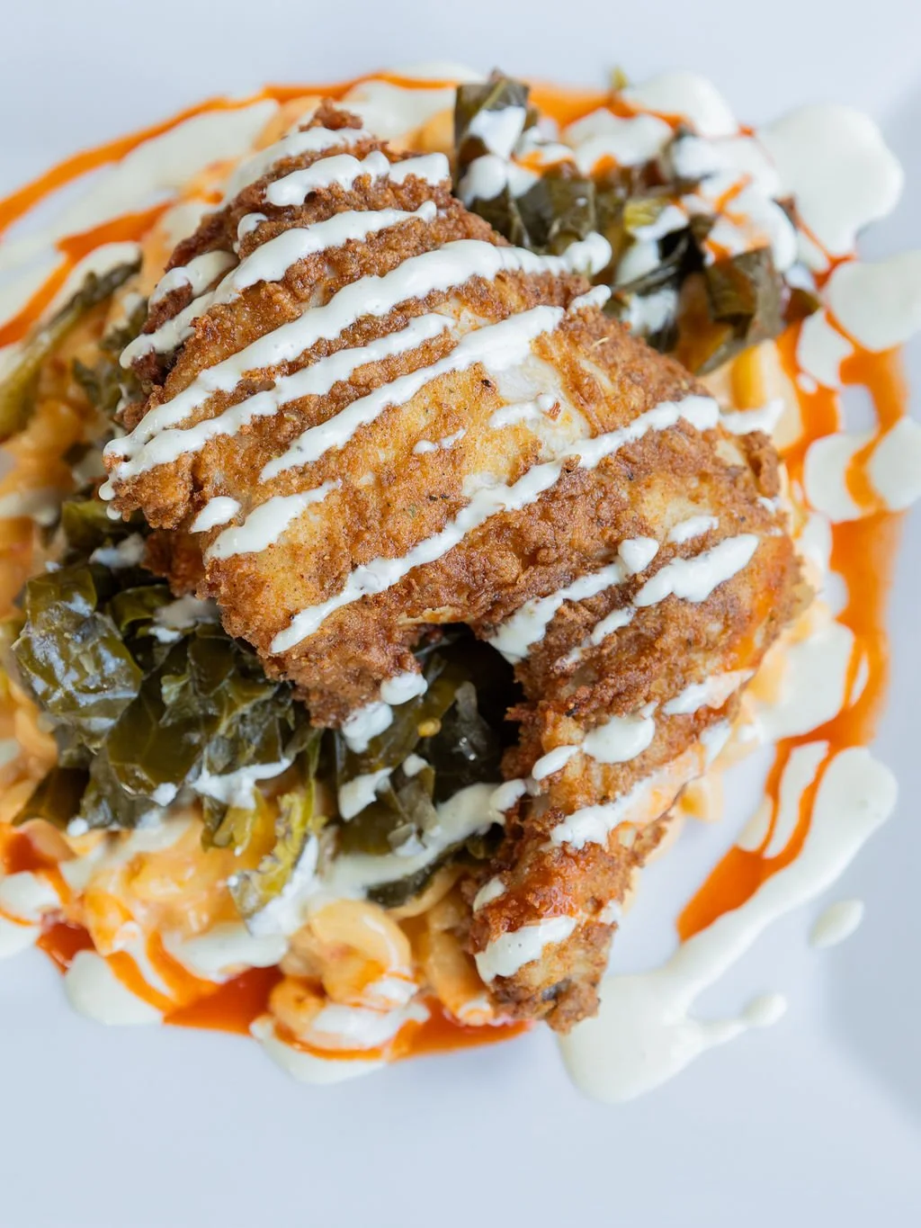 Fried fish fillet topped with drizzled white sauce, garnished with pickled greens, served over a bed of vegetables and sauce on a white plate.