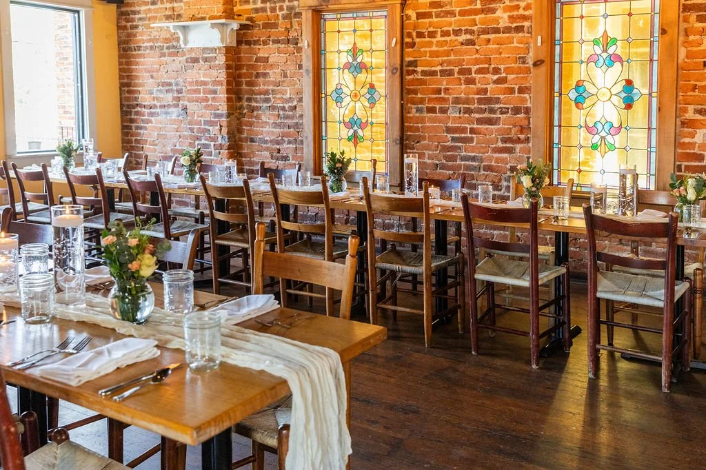 A restaurant dining area with wooden tables, flowers, candles, and stained glass windows, exposed brick walls, and natural light.