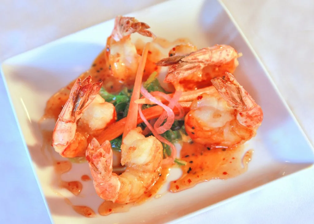 Four cooked shrimp with pink shells on a white square plate, garnished with green herbs and sliced vegetables, with sauce drizzled on and around them.