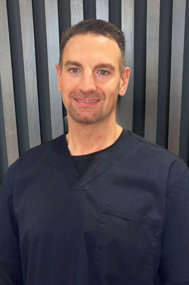 A smiling man in black medical scrubs standing in front of a modern wooden slat wall.