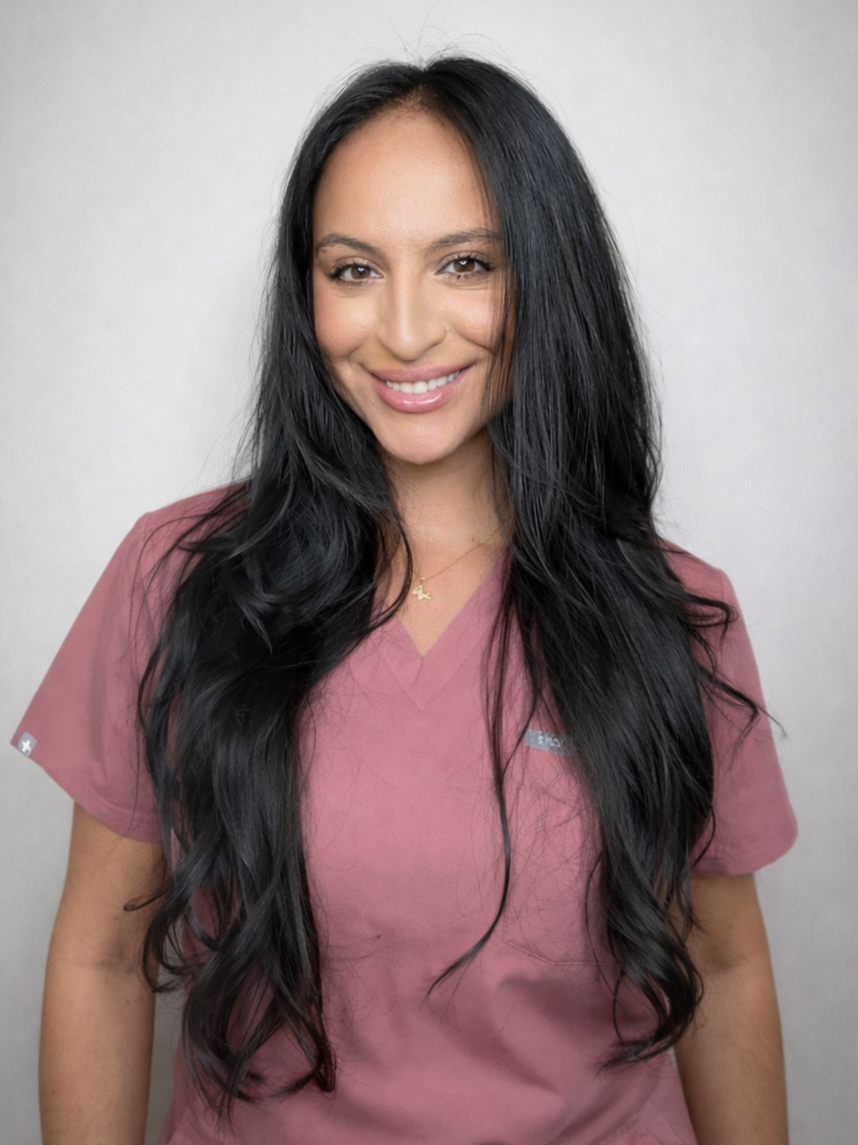 A smiling woman with long, wavy black hair wearing pink scrubs and a small necklace with a star pendant, standing against a plain light-colored background.