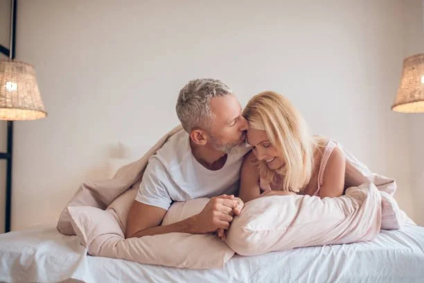 A couple sitting on a bed, the man kissing the woman on the forehead, both smiling and looking happy.
