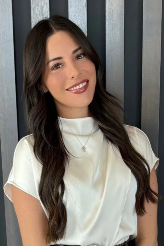 A woman with long dark hair and light skin, smiling, wearing a white silk blouse and a gold necklace with a star pendant, standing in front of a dark slat wall.