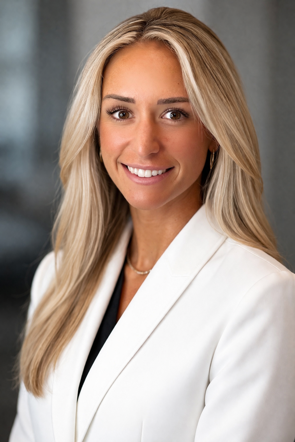 A professional woman with long blonde hair, smiling, wearing a white blazer and a necklace, in front of a neutral background.
