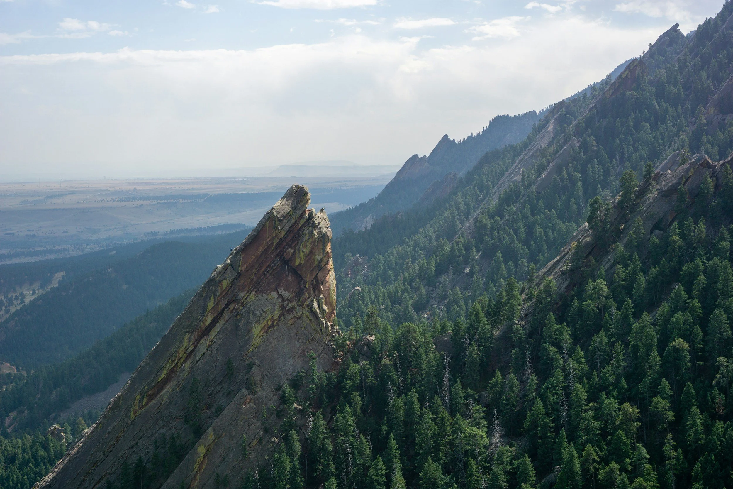 A mountain landscape with a prominent rocky peak surrounded by densely forested slopes and distant mountains under a partly cloudy sky.