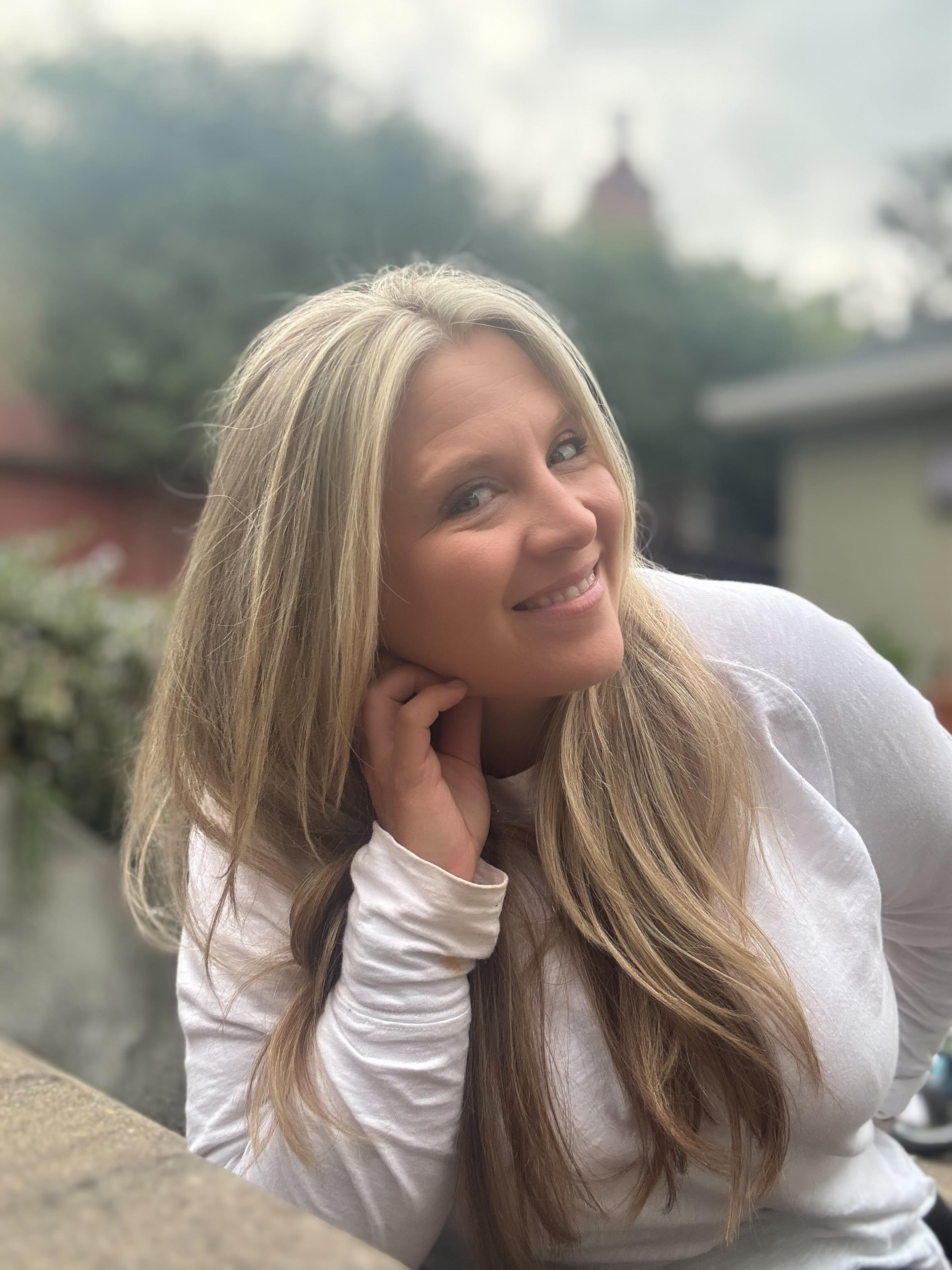 A woman with long blonde hair smiling at the camera, resting her head on her hand outdoors with trees and houses in the background.