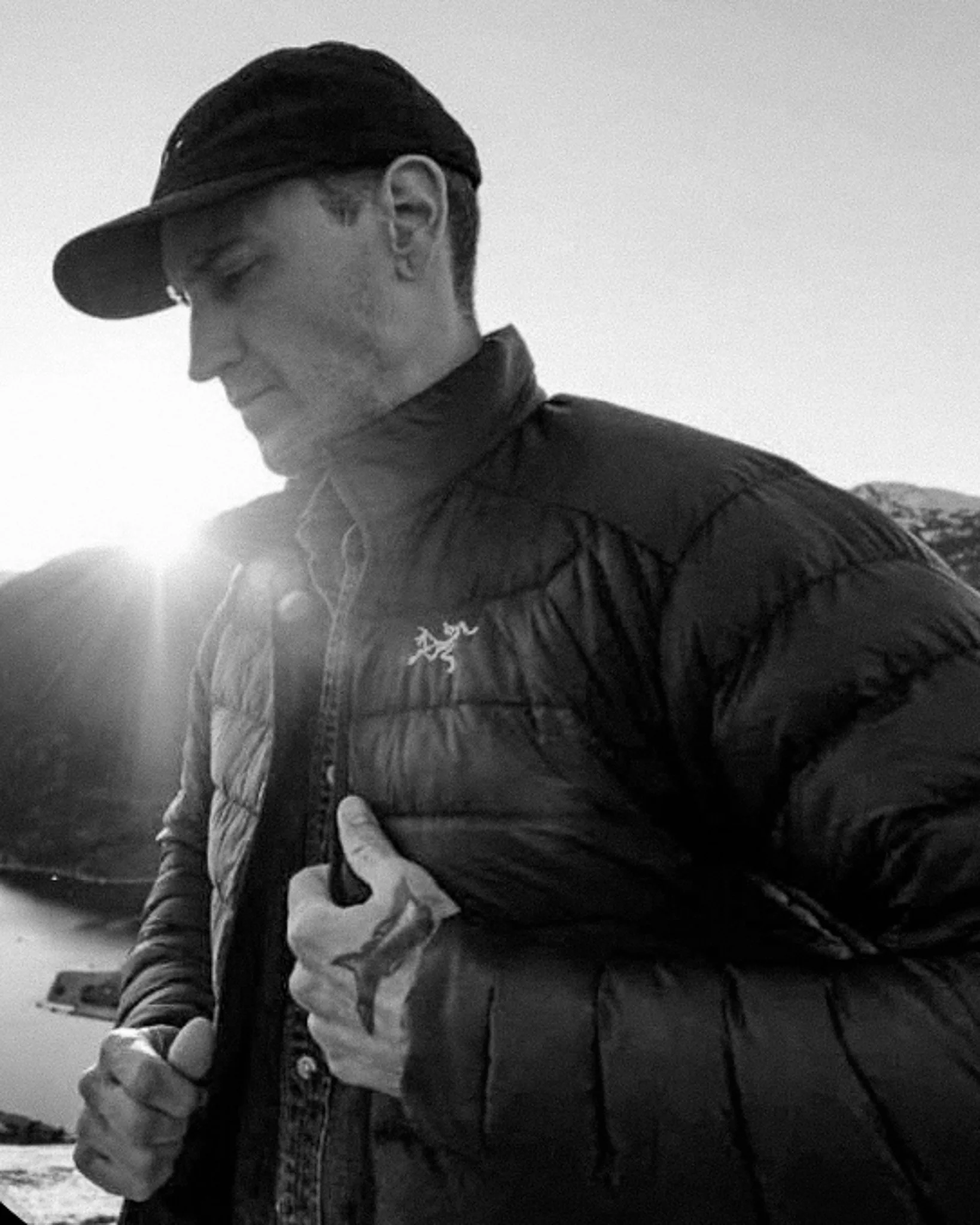 Black-and-white photo of a man wearing a cap and a puffy jacket, with his eyes closed, holding onto the zipper of his jacket, with a tattoo of a star on his hand, outdoors during sunset.