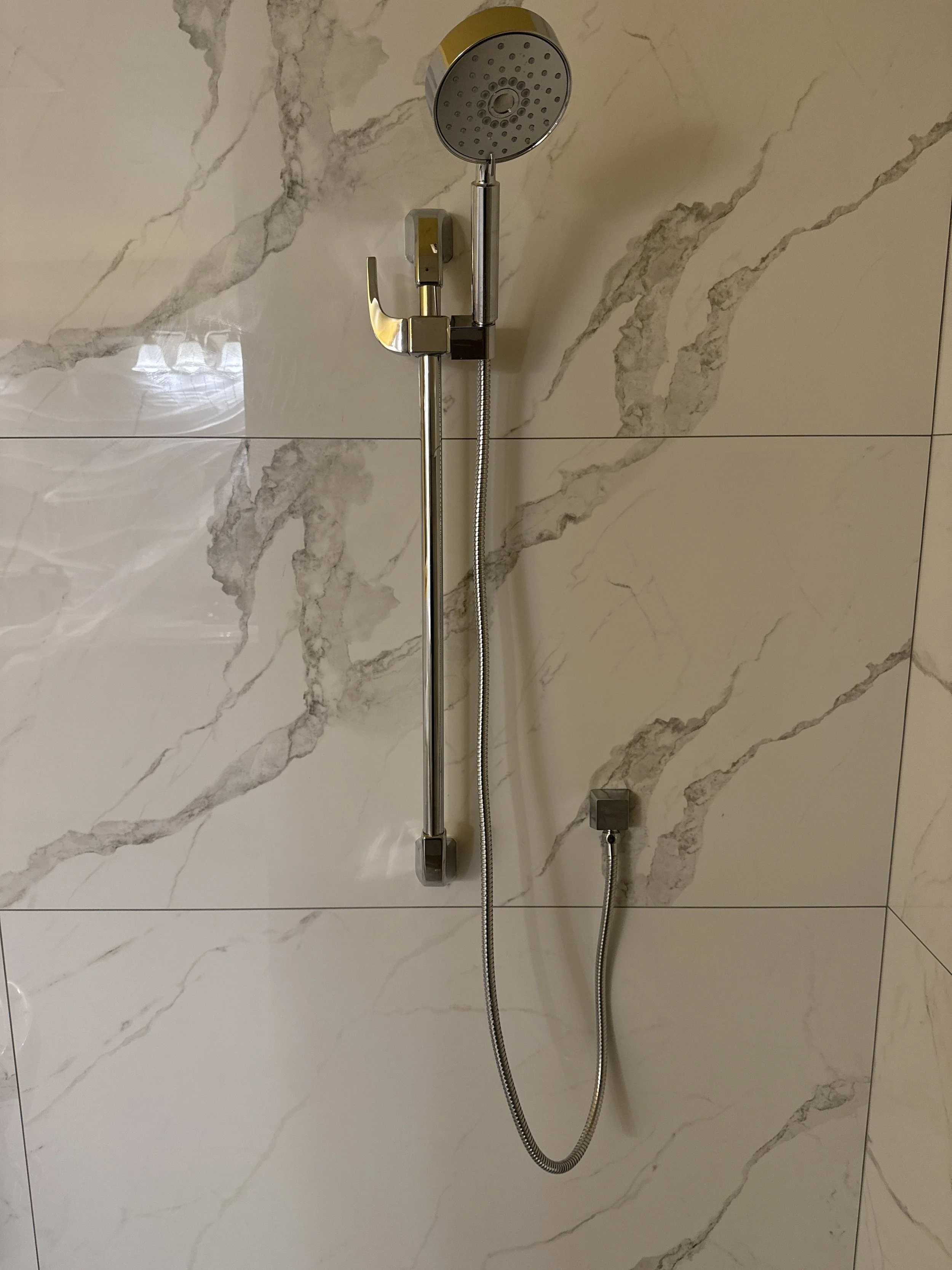 A showerhead with a handheld spray and wall mount, installed on a marble-tiled bathroom wall.