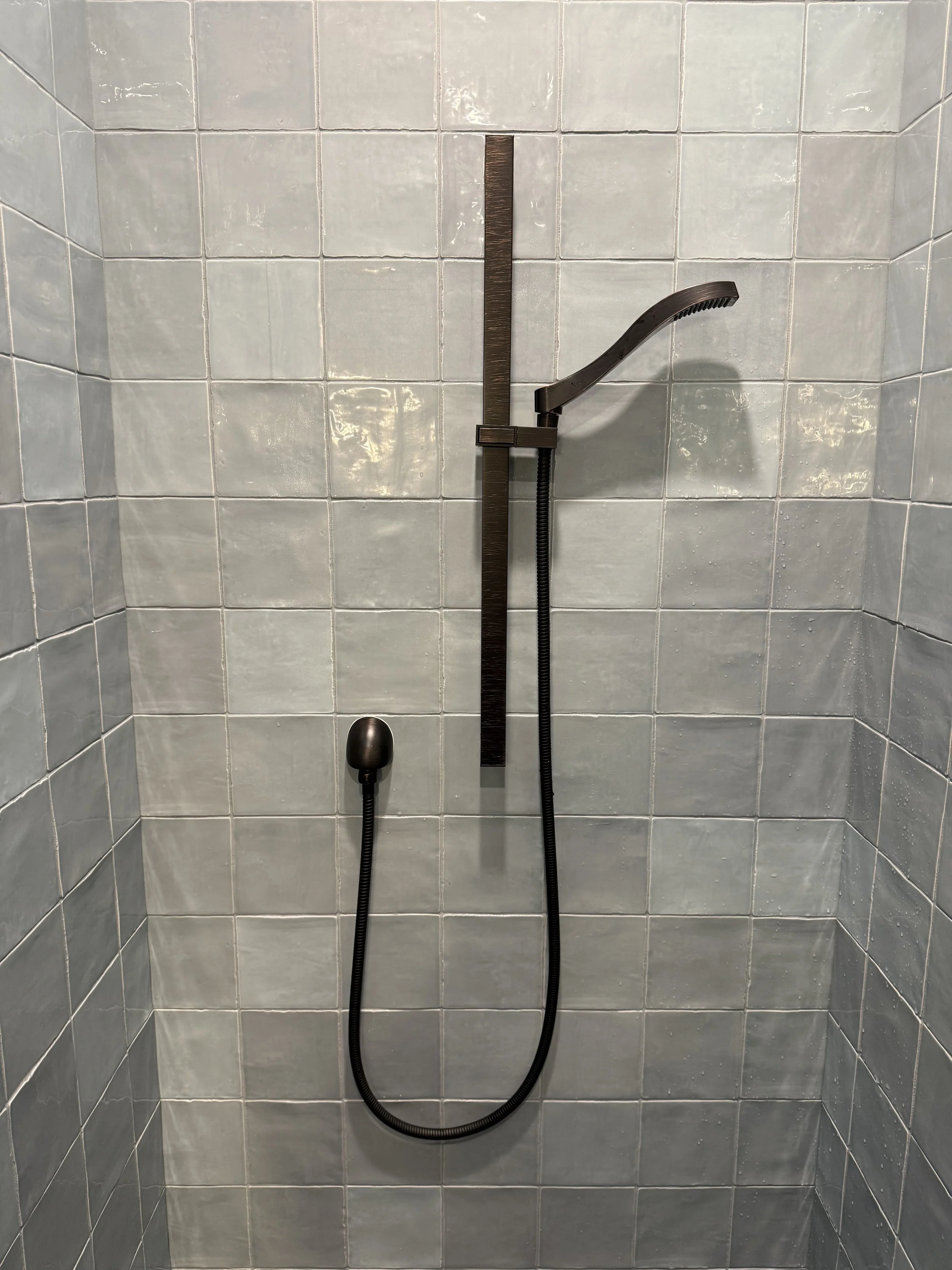 A shower with gray tiled walls and a black handheld showerhead attached to a vertical bar and hose.