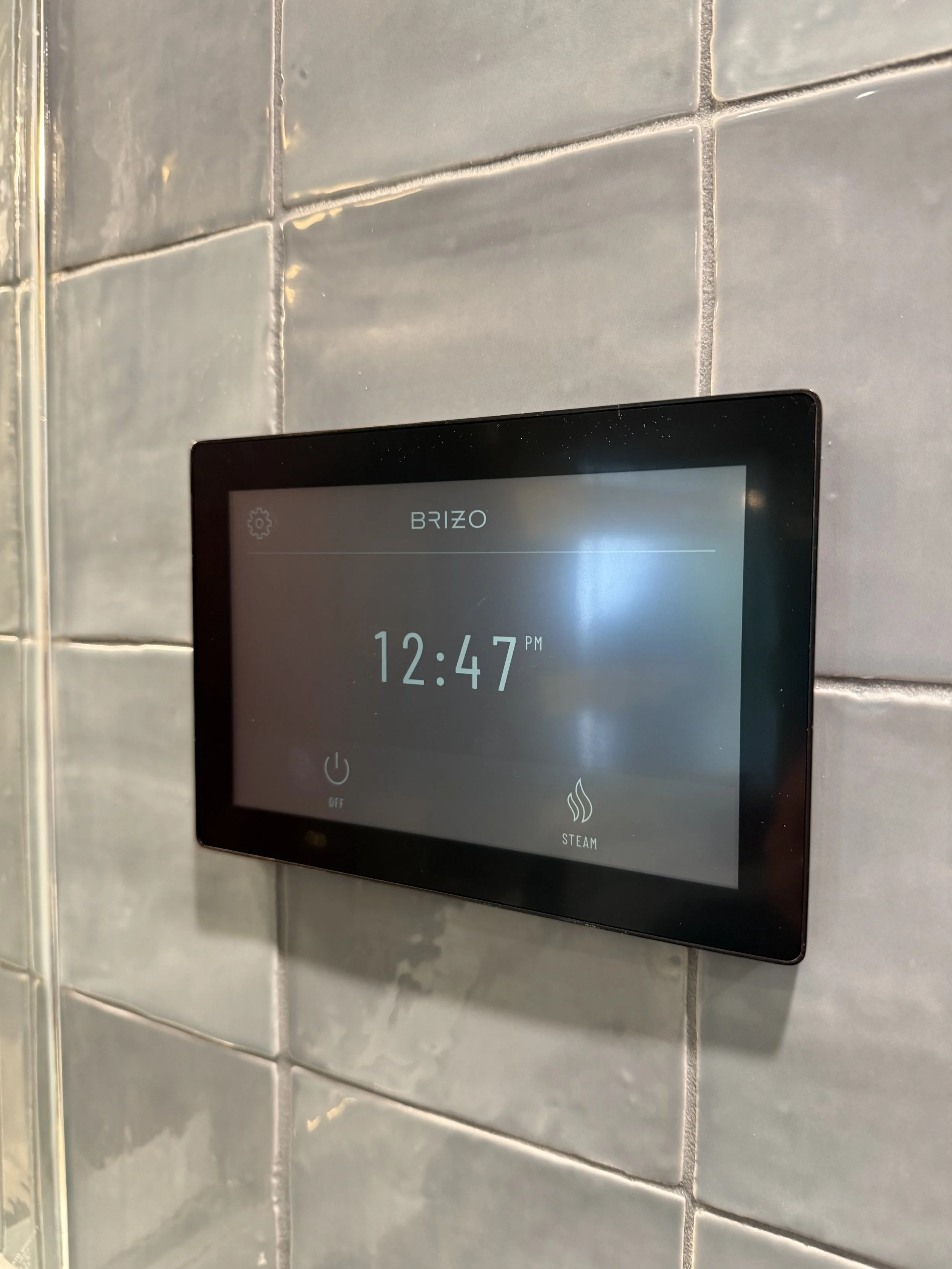 Digital control panel mounted on a tiled wall, displaying the time 12:47 PM, with icons for power off and steam functions.