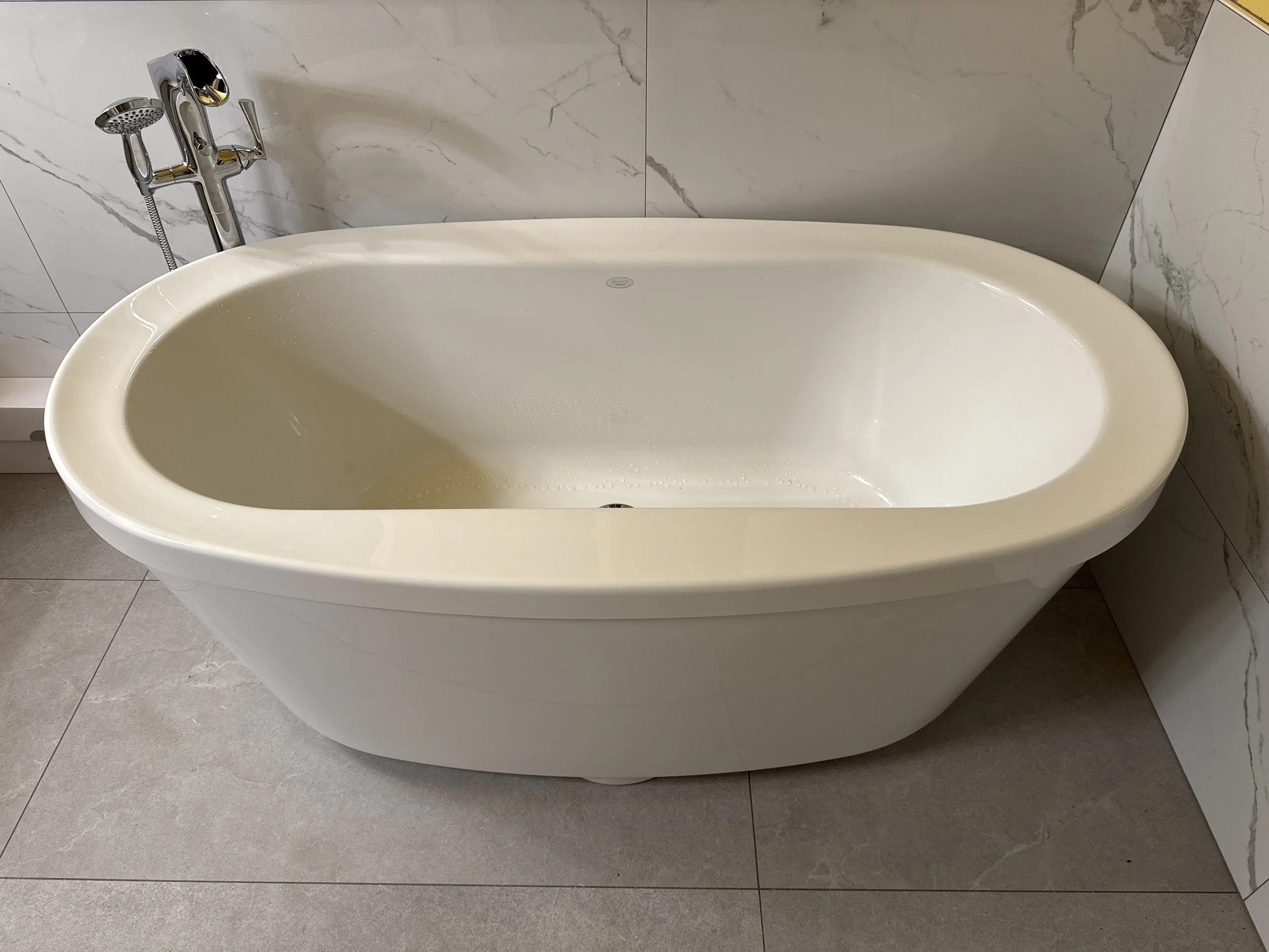 A modern, edge-free white freestanding bathtub in a bathroom with marble wall tiles and gray floor tiles.