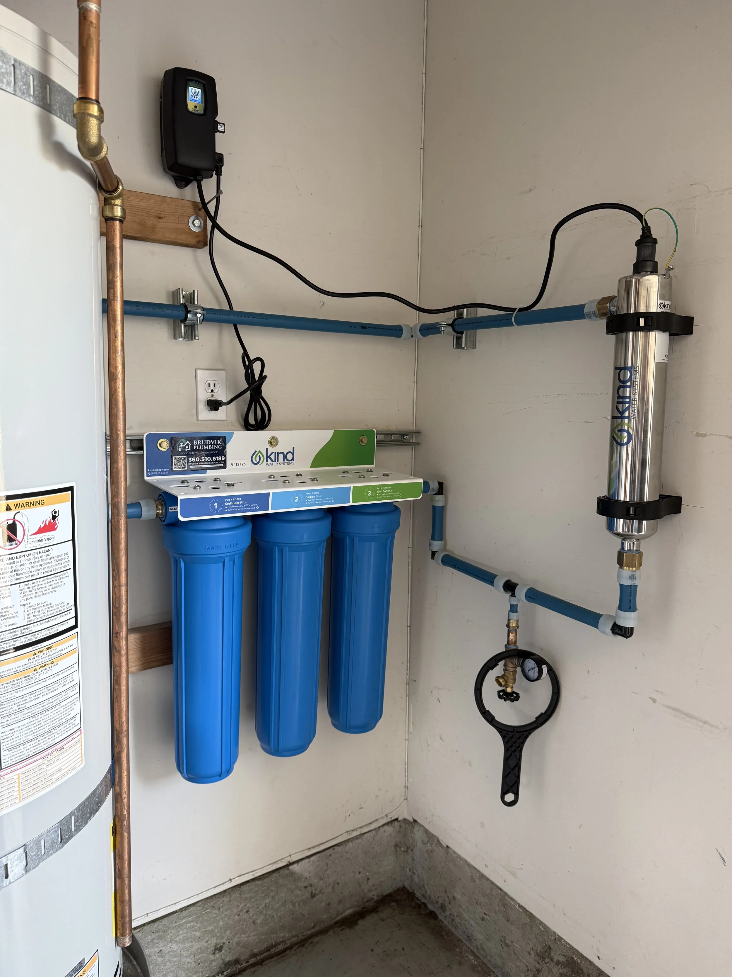 Under-sink water filtration system with three blue filter cartridges, connected pipes, a pressure gauge, and electronic water softener in a utility room.