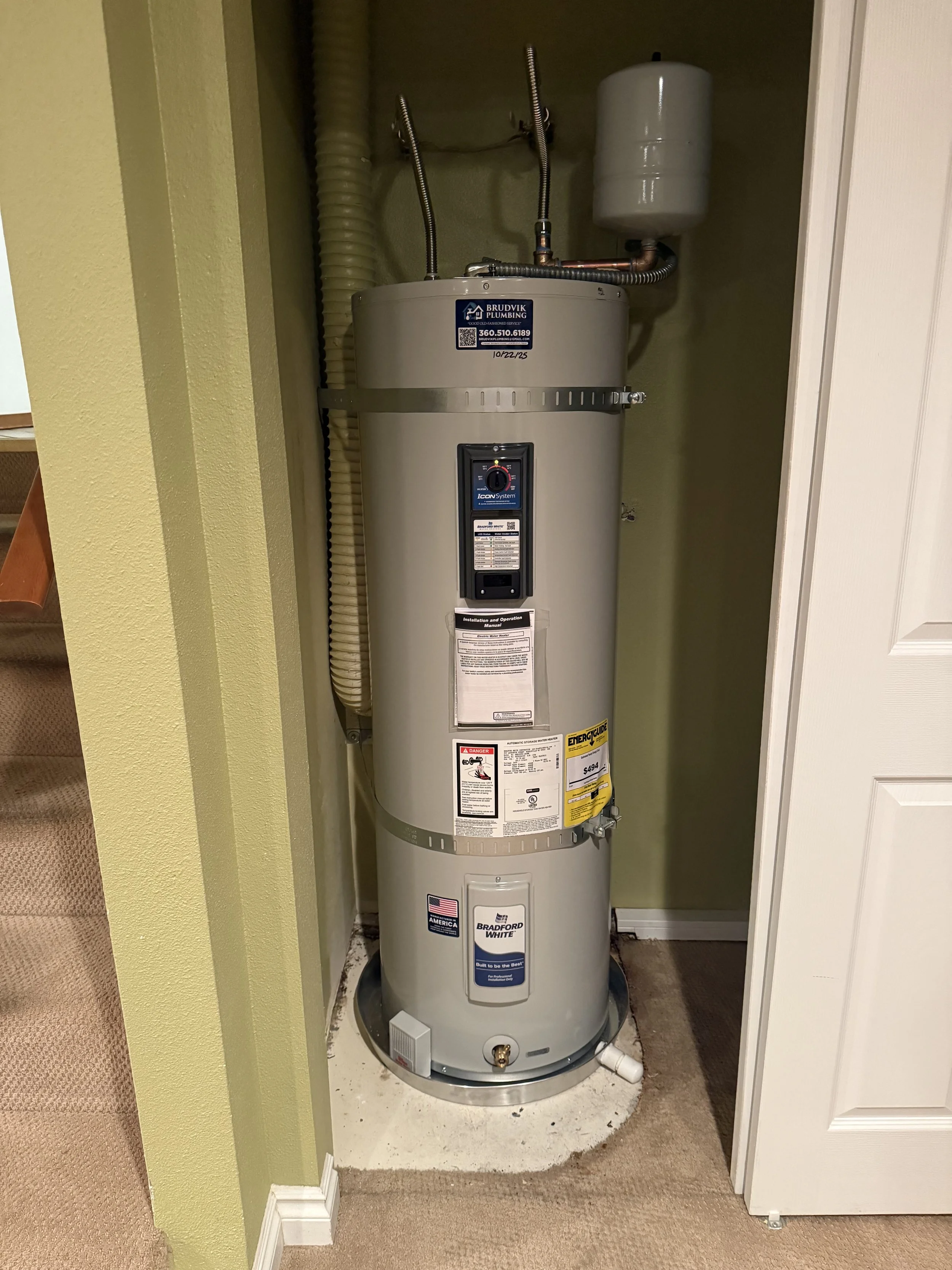 A tall, cylindrical water heater with various labels, pipes, and a pressure tank on top, situated in a small corner next to stairs and a white wall.