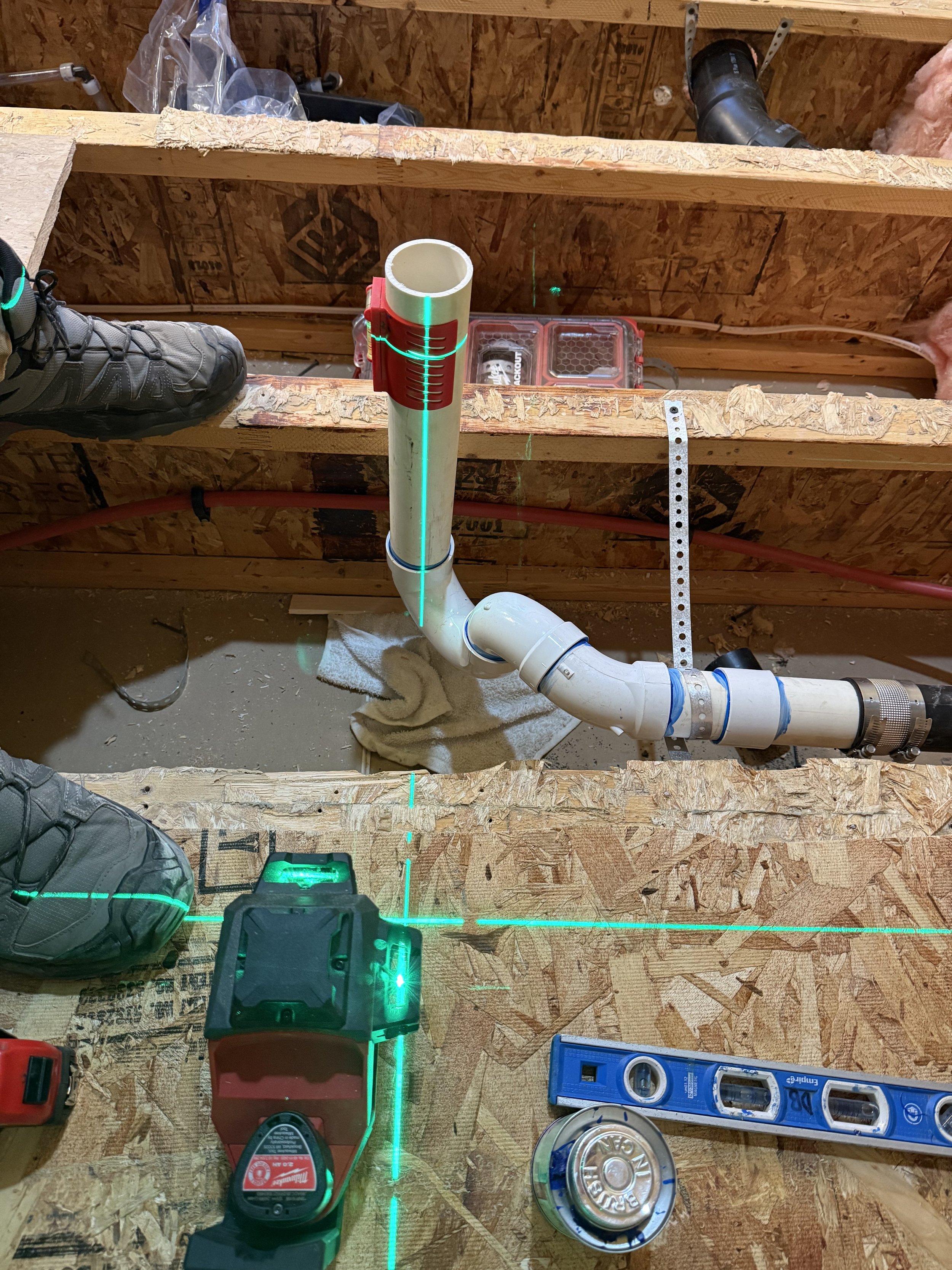 Construction site with plumbing pipes, a laser level projecting green lines, and construction tools on a plywood surface.