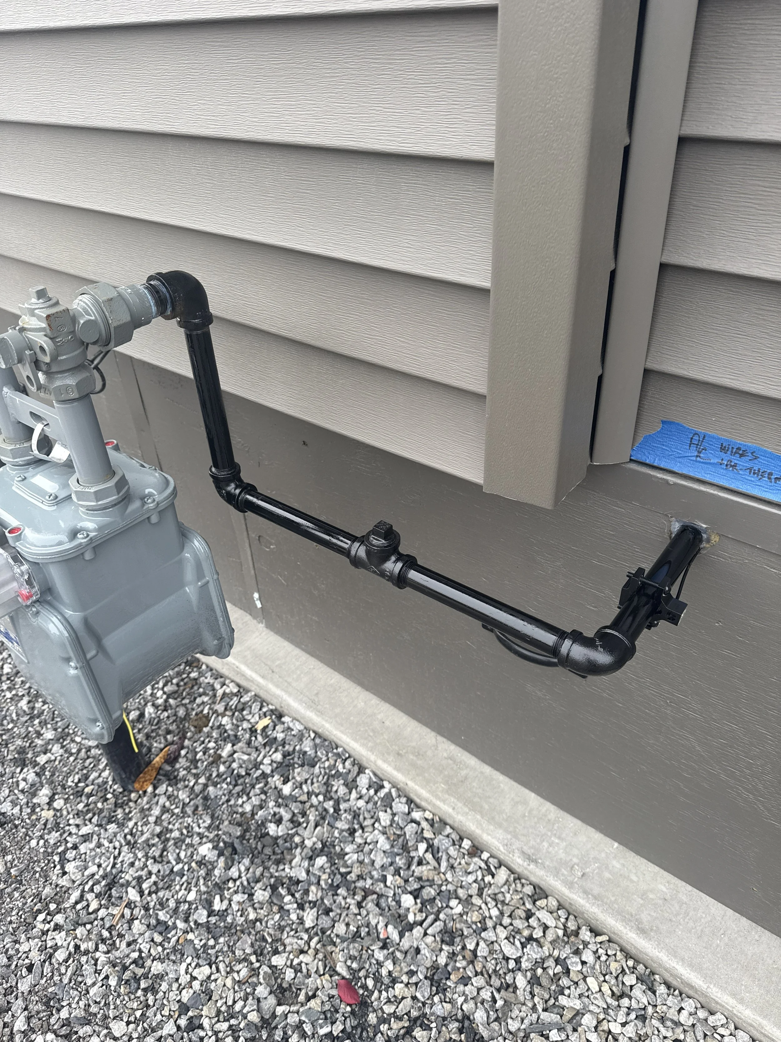 Gas meter connected to a black pipe running along the exterior wall of a house with vinyl siding, above a gravel ground surface.
