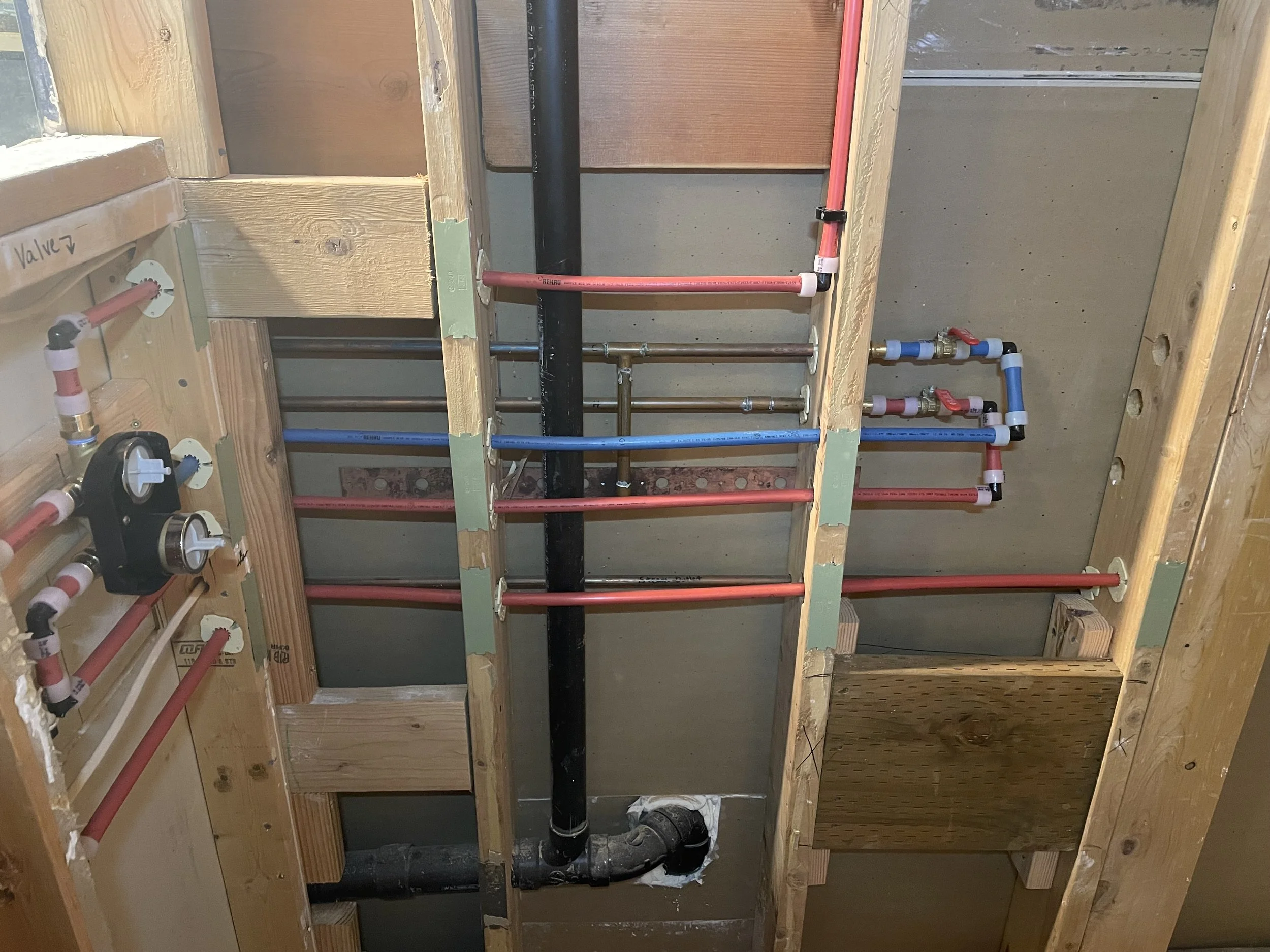 Interior of a building under construction showing plumbing with red and blue PEX pipes, black drain pipe, and wooden framing.