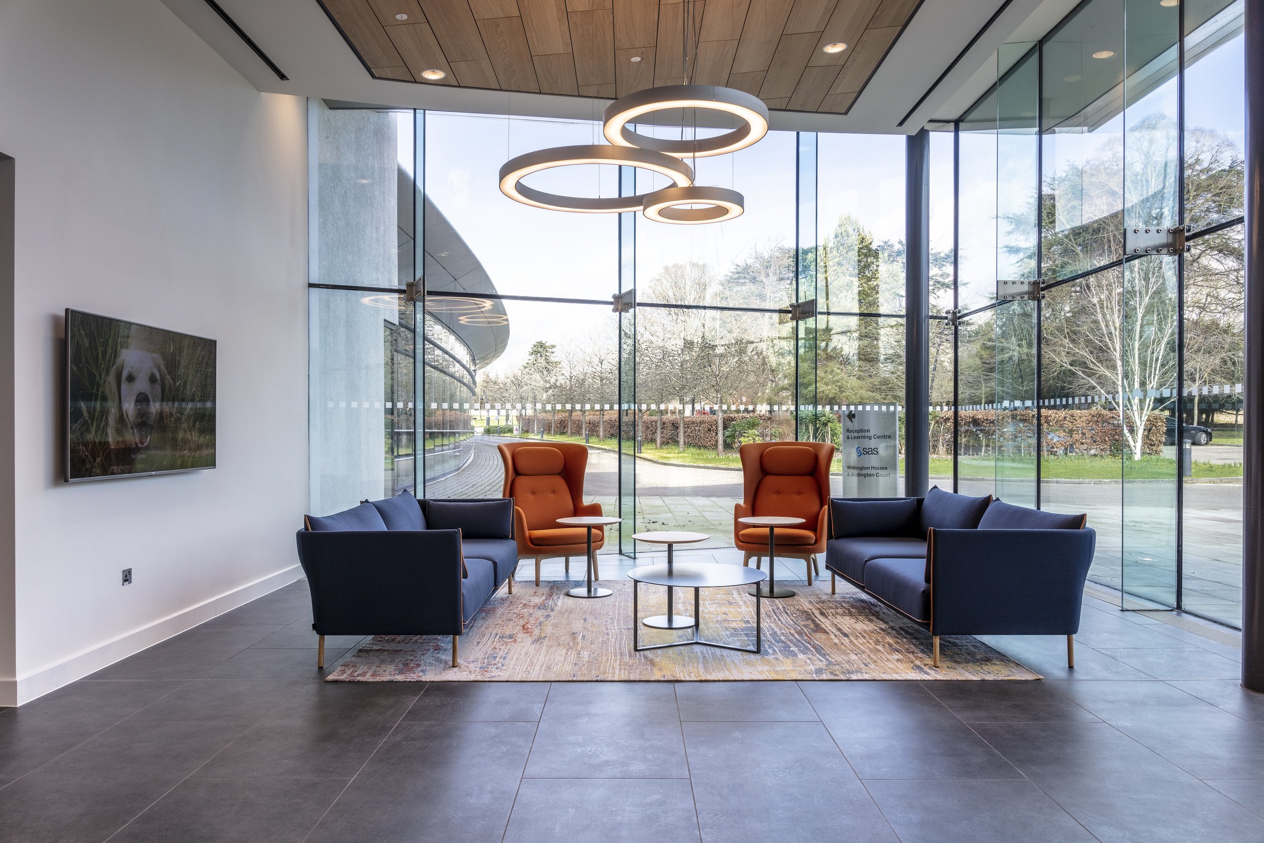 Modern reception area with large floor-to-ceiling windows, two dark blue sofas, two orange high-back chairs, multiple small tables, a colorful rug, and a circular ceiling light fixture.