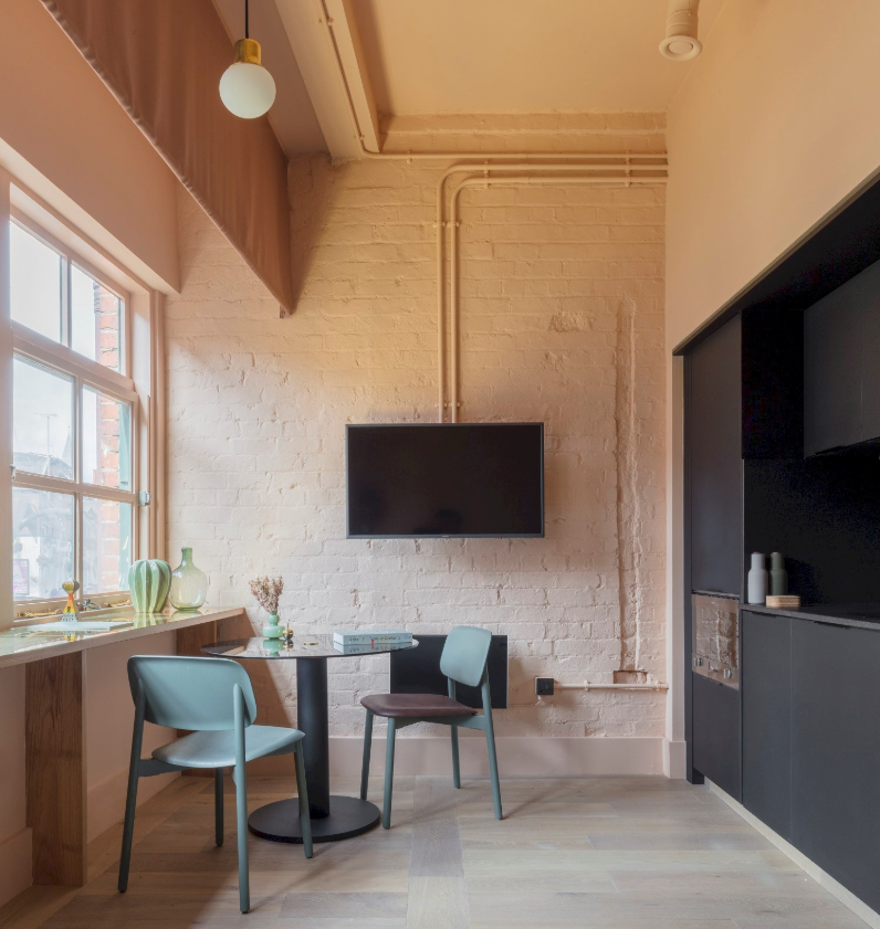 A cozy room with a white brick wall, a mounted flat-screen TV, a small round dining table, two chairs, a window with a view outside, and a black cabinet with decorative items.