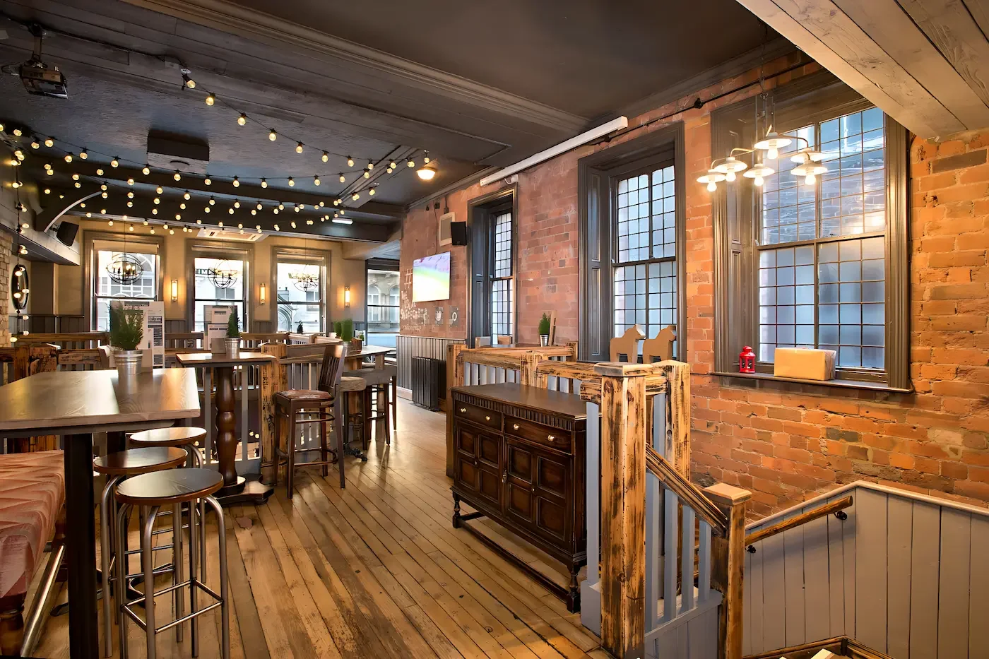 Interior of a cozy restaurant or pub with exposed brick walls, wooden furniture, large windows, string lights on the ceiling, and modern light fixtures.