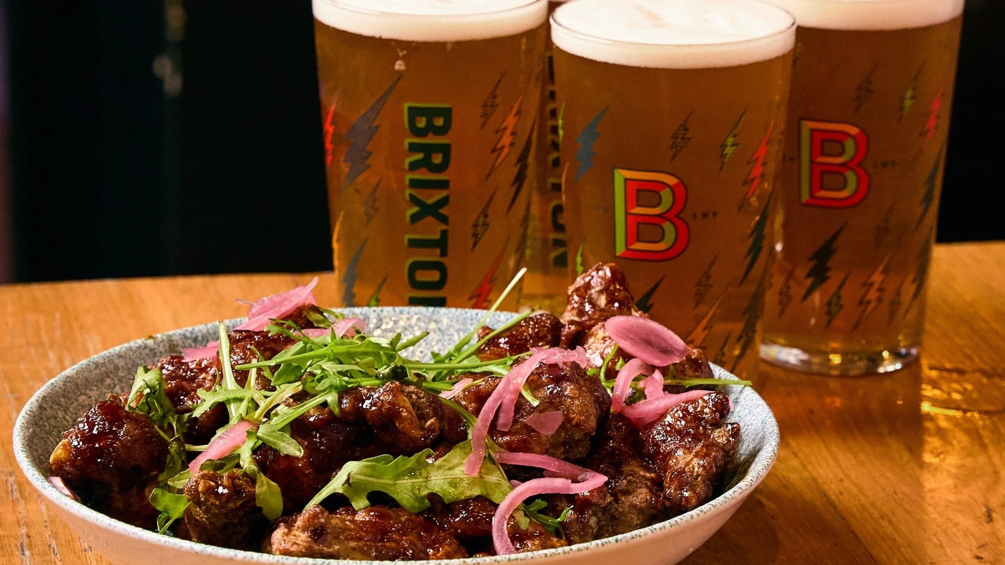 A bowl of glazed chicken wings garnished with greens and pickled onions, with three glasses of light beer in the background on a wooden table.