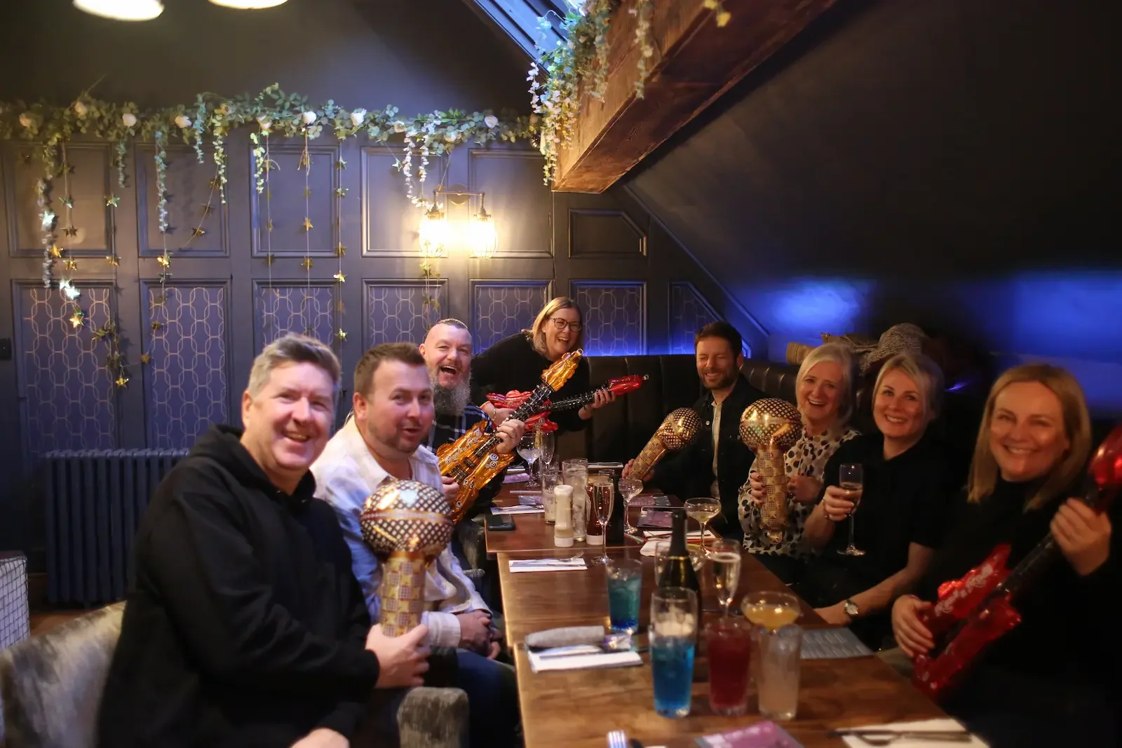 Group of people celebrating at a dinner party, holding colorful guitar-shaped and orb-shaped decorative objects, with smiles, drinks, and an evening ambiance in a decorated restaurant.