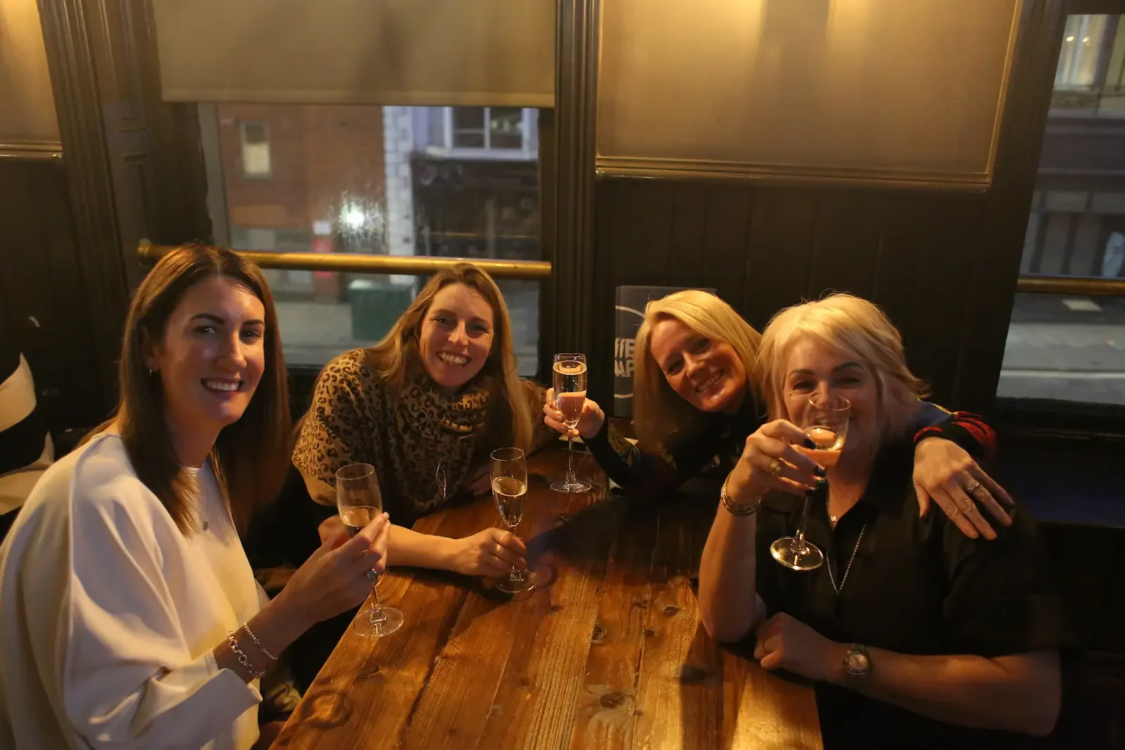 Four women sitting at a wooden table in a bar or restaurant, smiling and holding glasses of Champagne or sparkling wine, celebrating together.