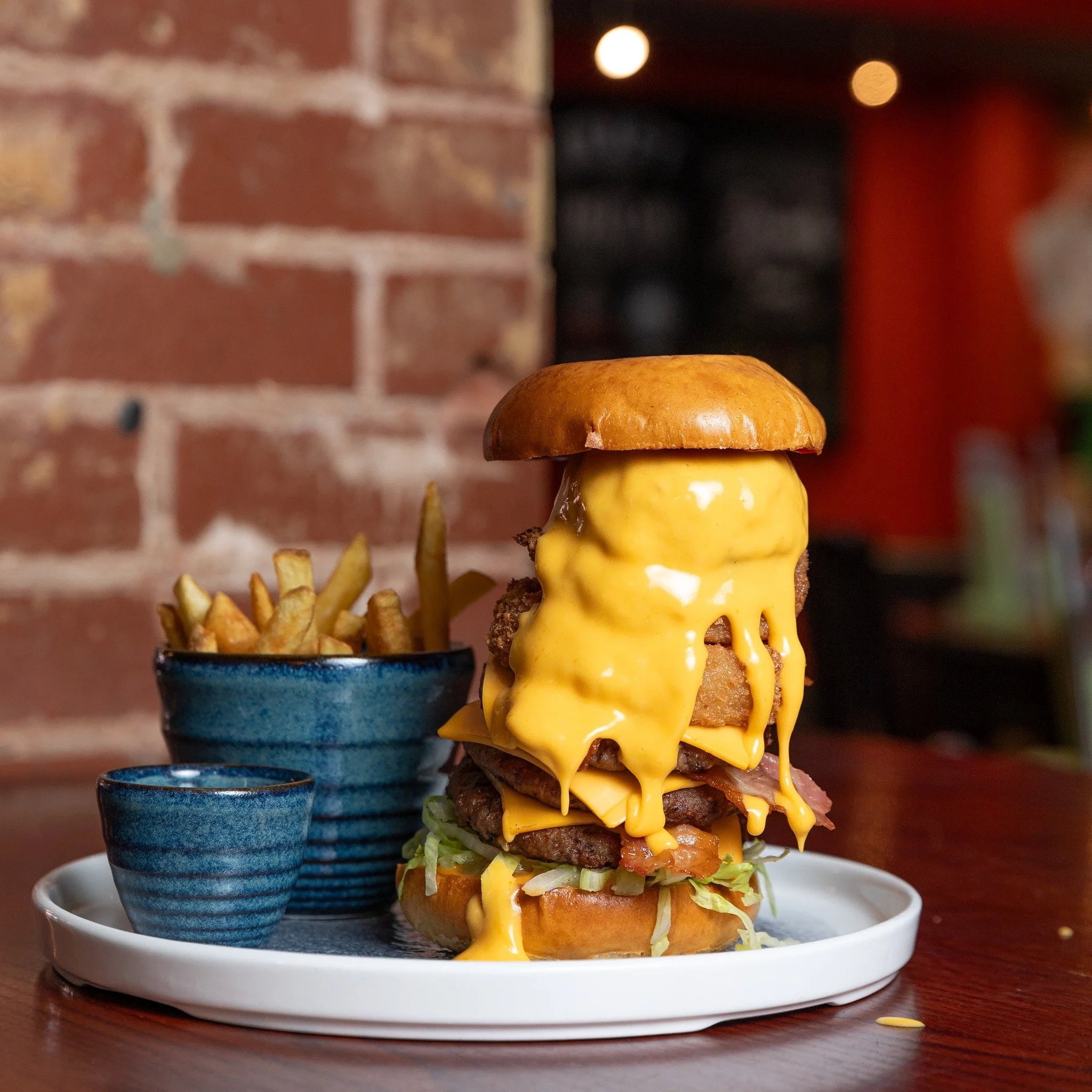A towering cheeseburger with multiple patties, melted cheese, lettuce, bacon, and onion rings, served with French fries in a blue bowl and a small blue cup on a white tray in a restaurant setting.