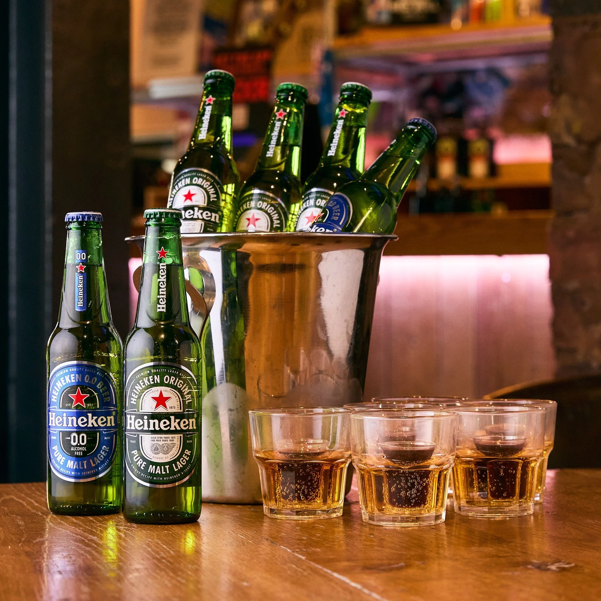 Six bottles of Heineken beer, three bottles of Heineken 0.0, and four glasses of beer on a wooden table in a bar setting with a metal bucket and a brick wall in the background.