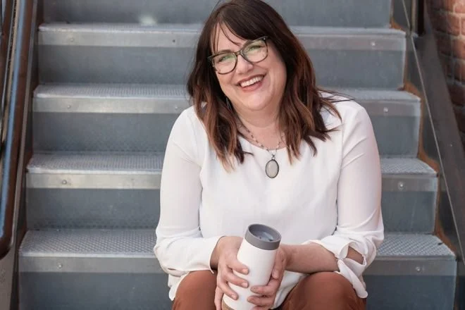 A woman with glasses and shoulder-length brown hair sitting on metal stairs, smiling, holding a white tumbler.