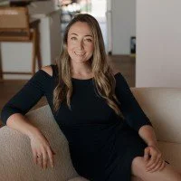 Woman with long brown hair wearing a black dress sitting on a beige couch in a room with wooden furniture and a lamp