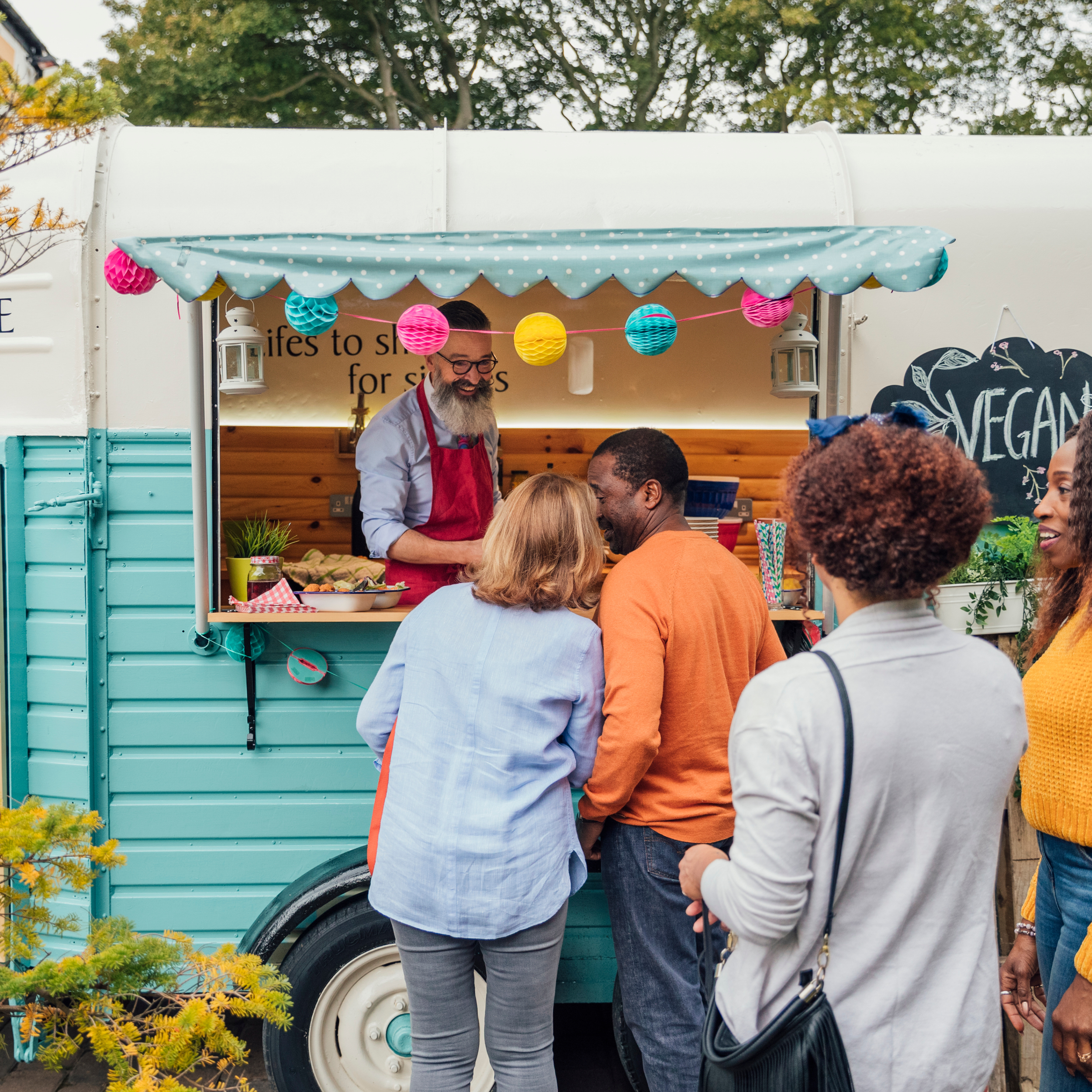 A group of people, including a woman with blonde hair in a light blue blazer and a man in an orange shirt, standing in front of a teal and white vegan food truck decorated with colorful paper lanterns. Inside the truck, a man with glasses and a beard, wearing a red apron, is preparing food, with a sign behind him reading 'lives to sh for si' and a chalkboard sign saying 'VEGAN' with a leafy design.