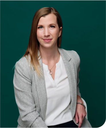 A woman with shoulder-length light brown hair wearing a gray blazer, white blouse, and a silver necklace, standing in front of a green background.