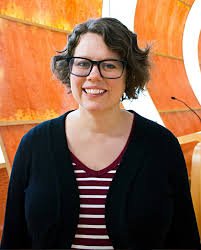 Woman with short curly hair, glasses, wearing a black cardigan over a red and white striped shirt, standing in front of an orange and white abstract background.