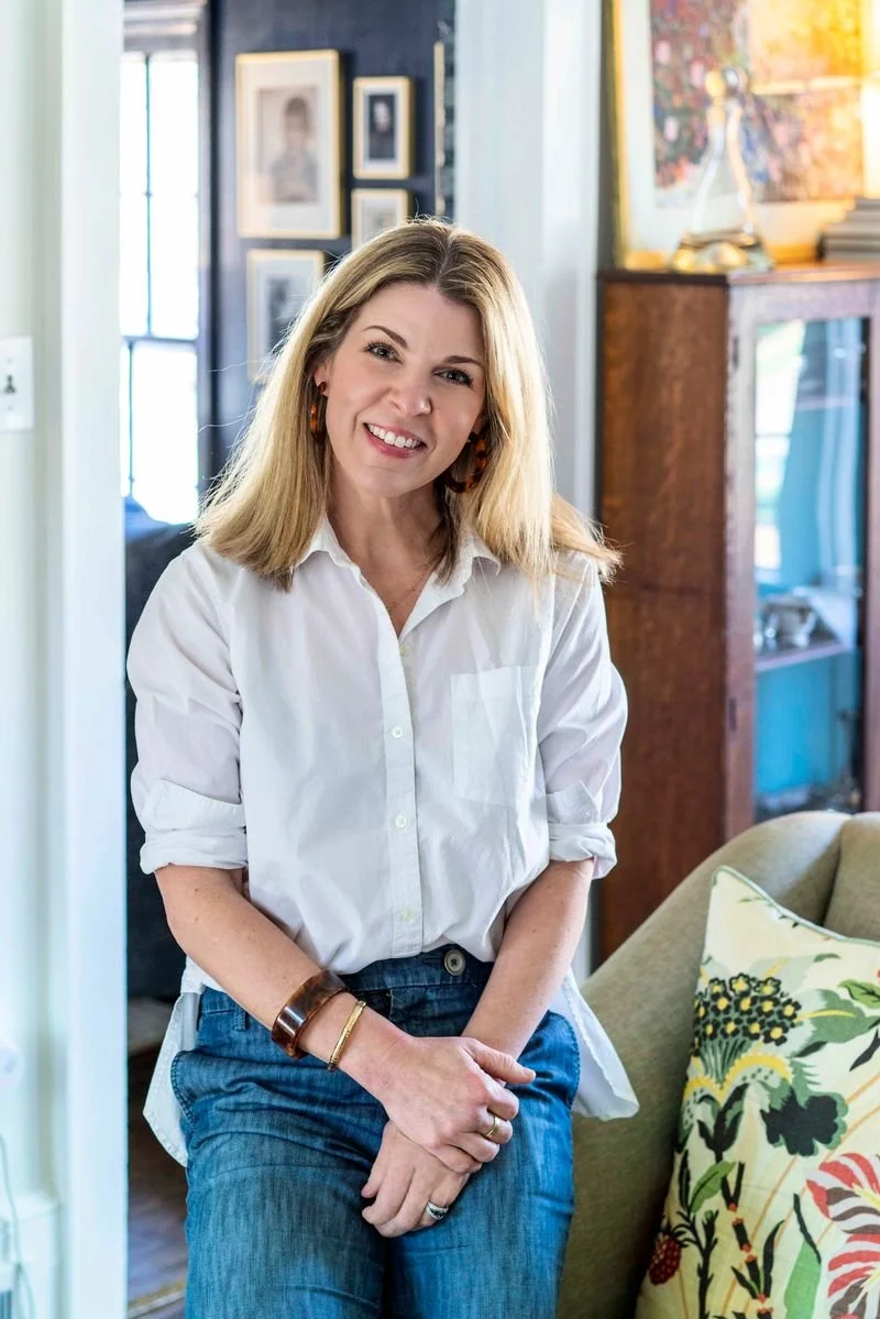 A woman with blonde hair wearing a white shirt and blue jeans, smiling and standing in a cozy living room.
