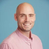 Portrait of a bald man with a light pink shirt smiling against a light blue background.