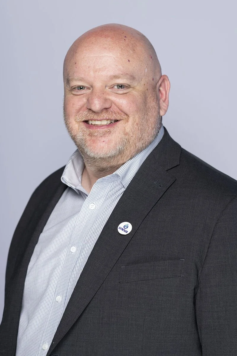 A smiling man with a shaved head and beard, wearing a dark suit and a light-colored collared shirt, standing against a plain background.
