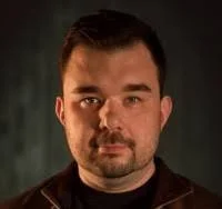 A portrait of a man with short dark hair and a beard, wearing a dark shirt, against a dark background.