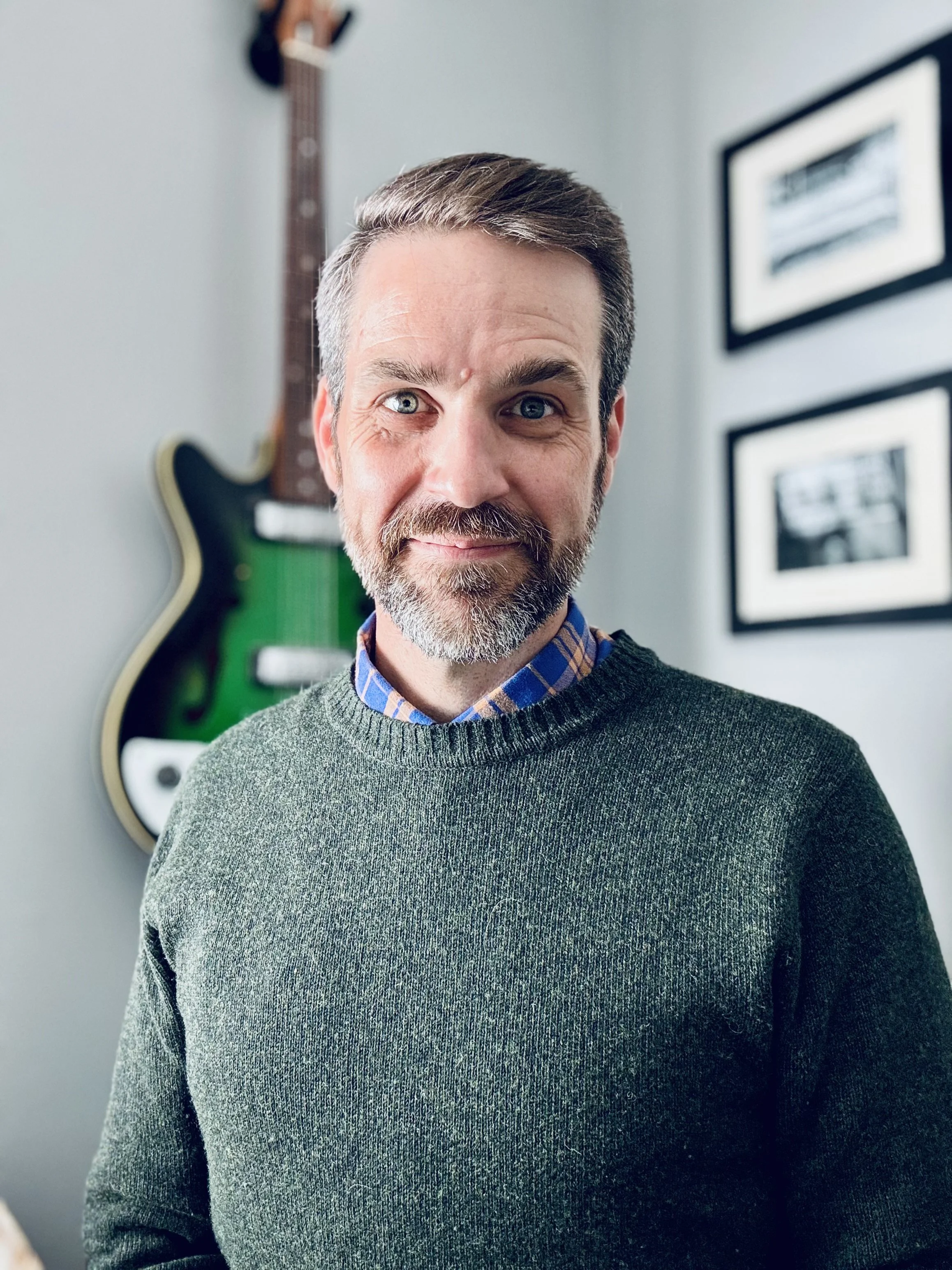A man with a beard and mustache, with one eye slightly squinted, standing indoors against a wall with framed pictures and a guitar hanging in the background.