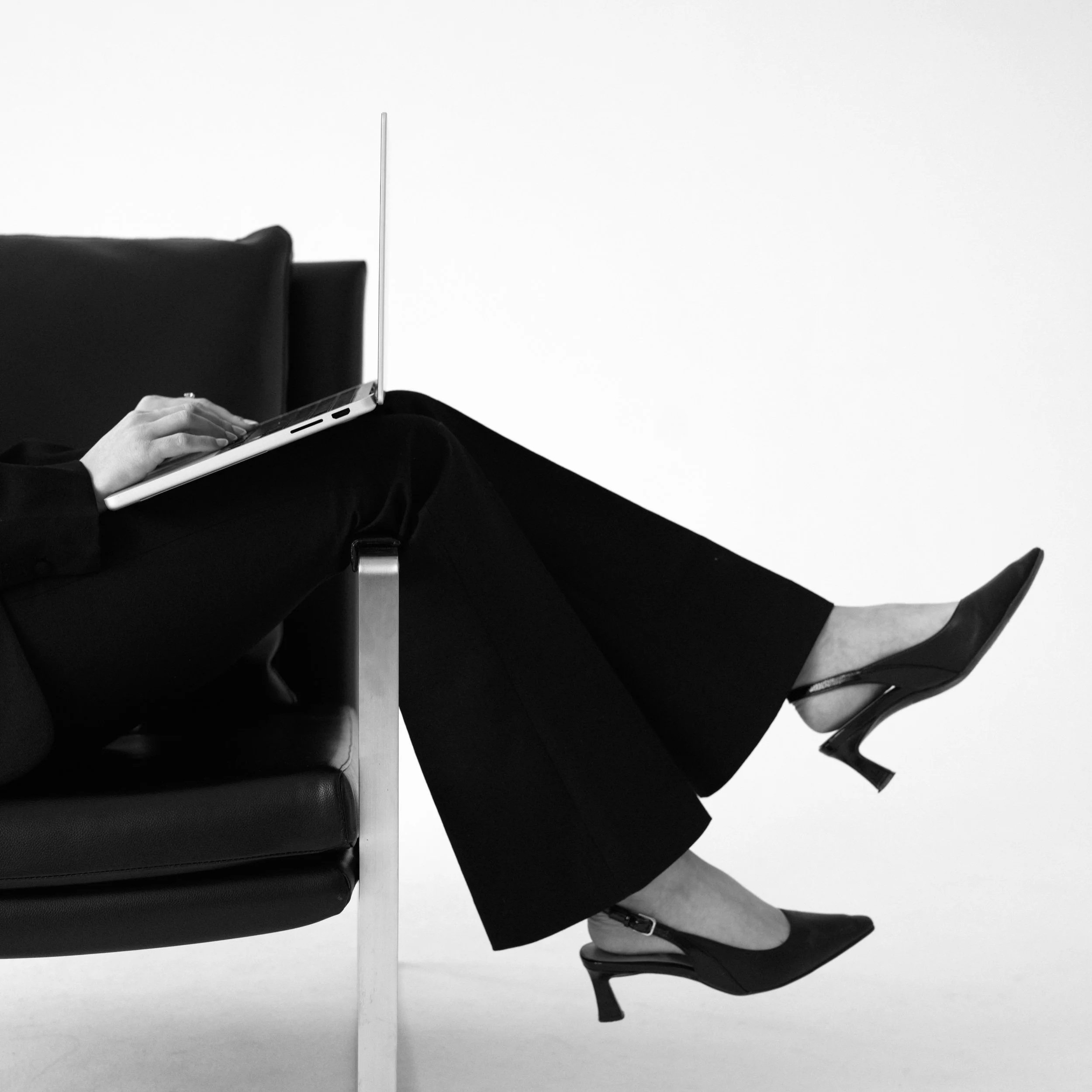 Black and white photo of a woman sitting on a modern armchair, wearing black pants and black high heels, working on a laptop with her legs extended.