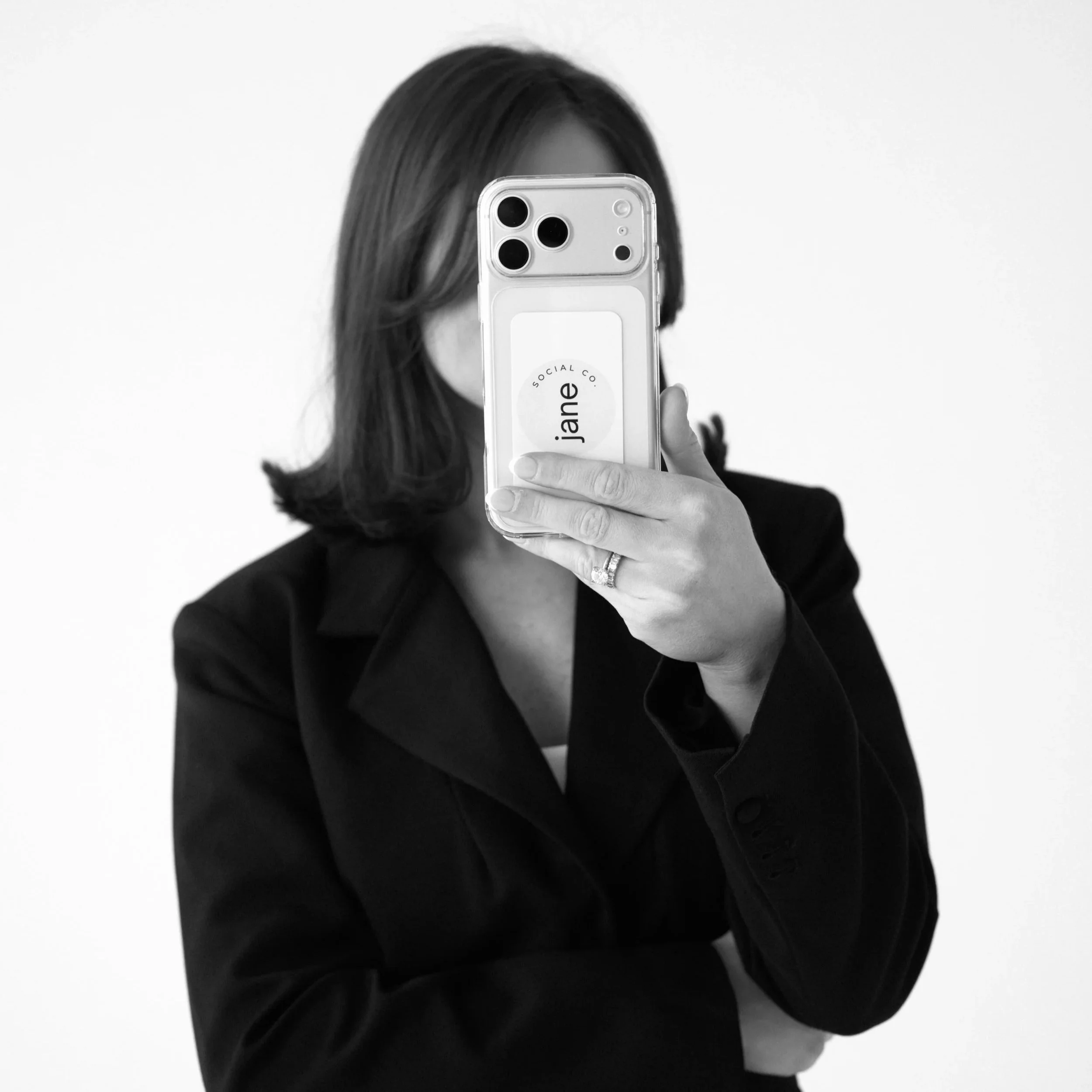 A woman with shoulder-length hair wearing a black blazer taking a selfie with her smartphone, obscuring her face.