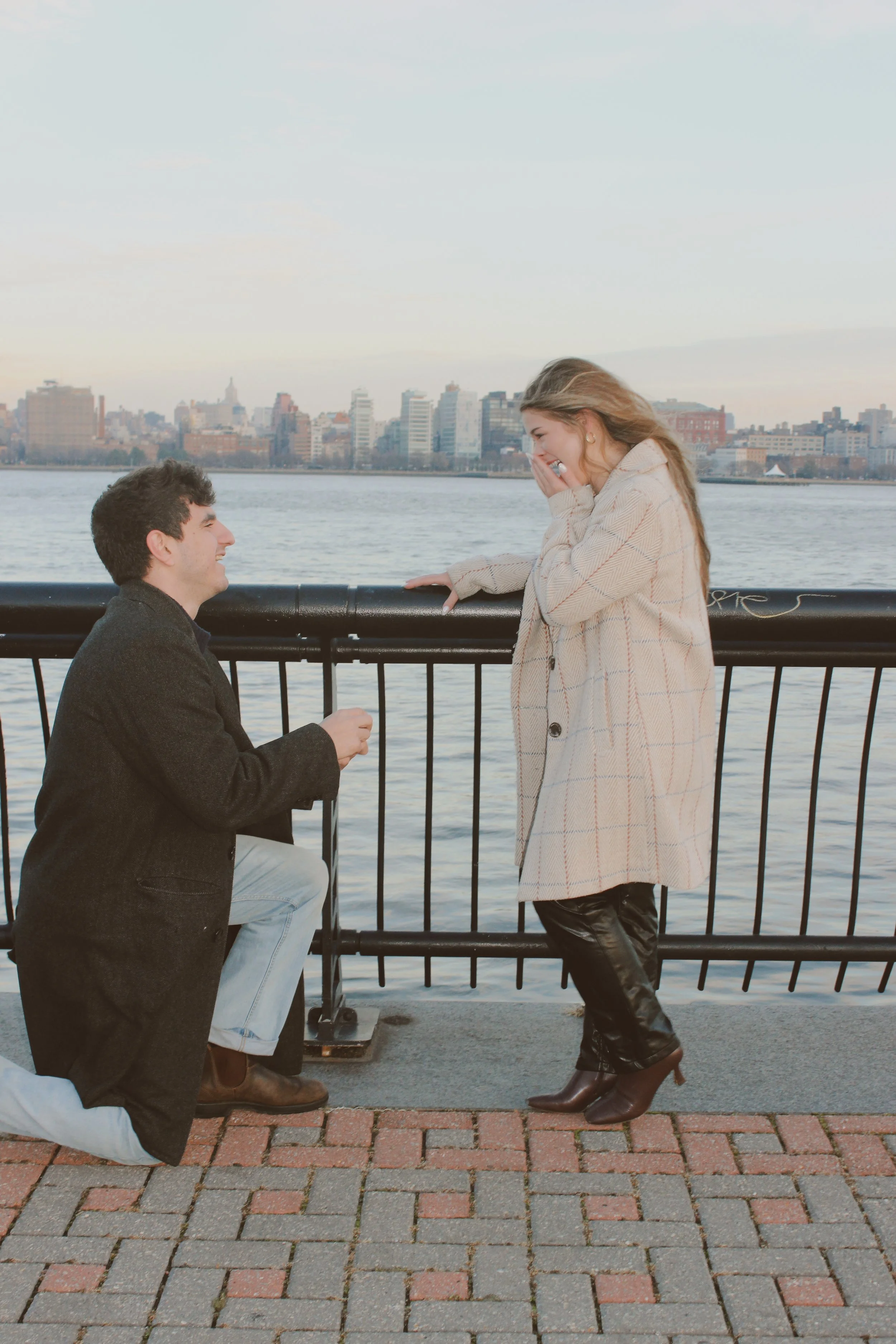 A man proposing to a woman by a river with the city skyline in the background. The man is kneeling on one knee, holding a small box, while the woman stands with her hands covering her mouth, smiling happily.