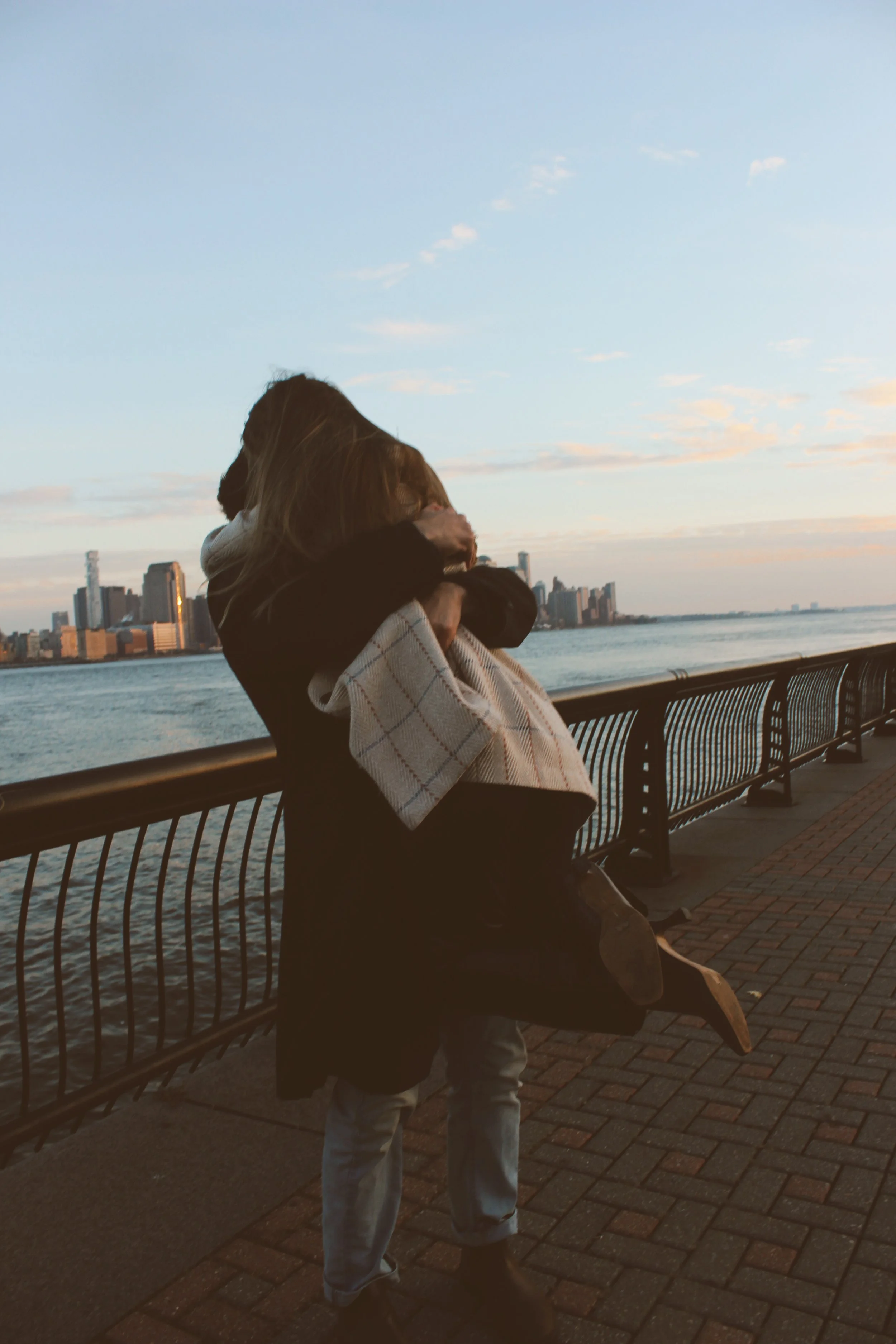 Two people hugging each other on a waterfront promenade during sunset with a city skyline in the background.
