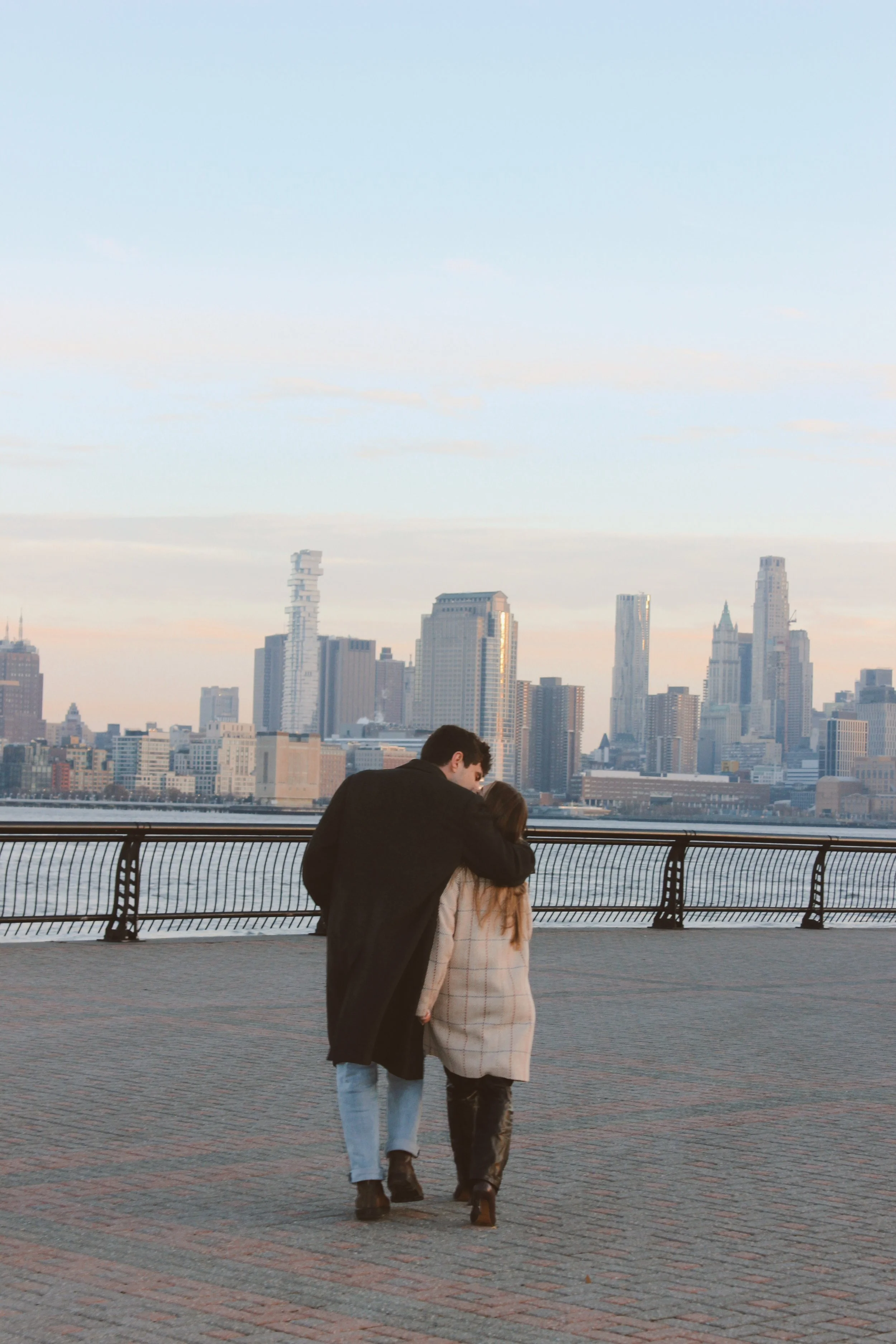 A young couple walking arm-in-arm along a waterfront promenade with a city skyline in the background during dusk.