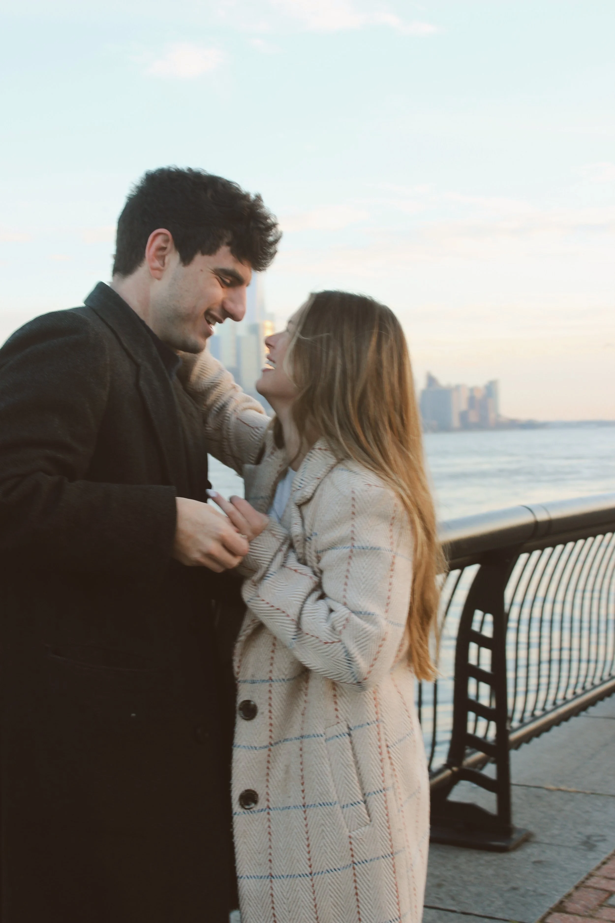 A couple smiling and laughing together by a waterfront, with city buildings in the background during sunset.