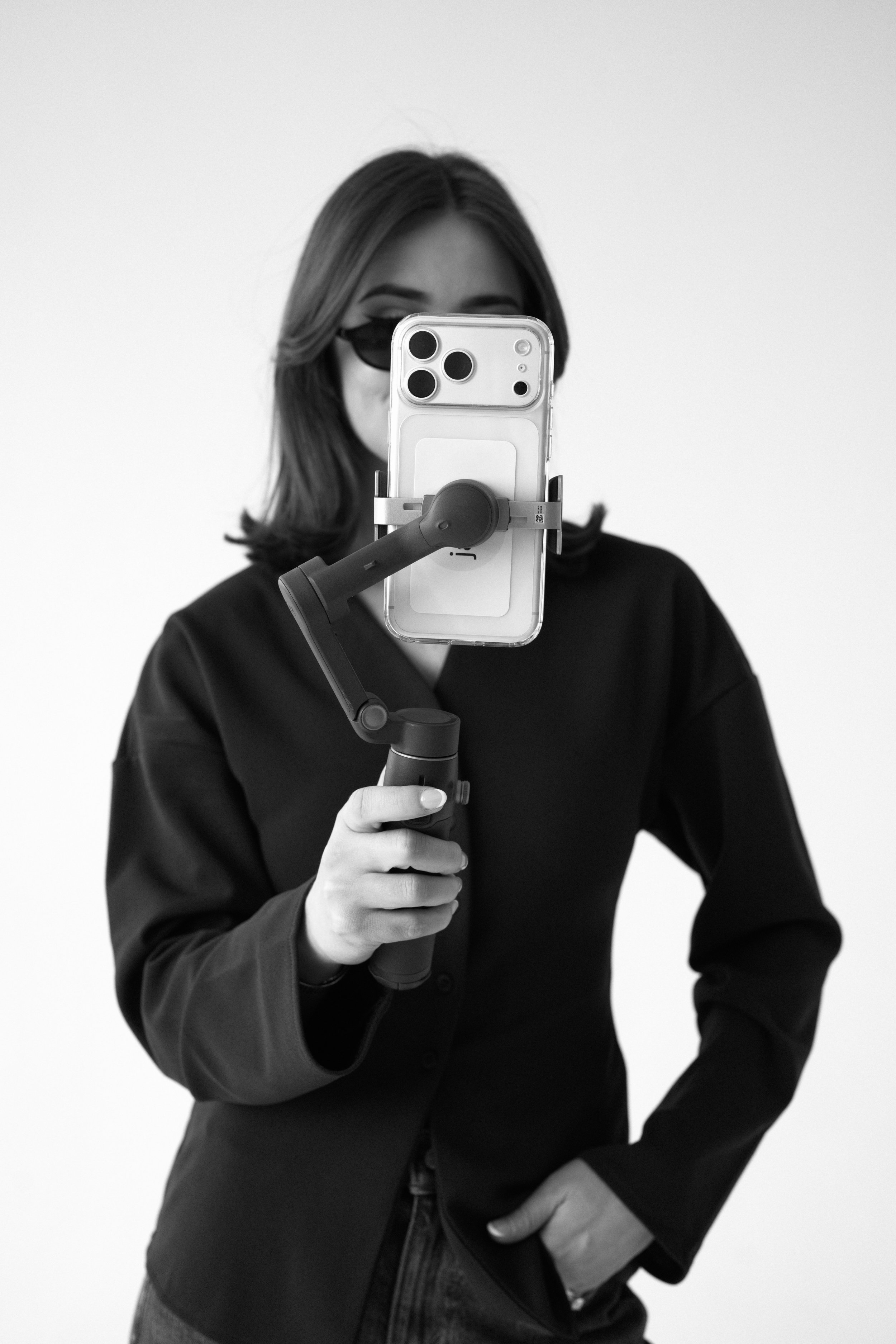 A woman taking a selfie with a phone attached to a handheld stabilizer against a plain background.