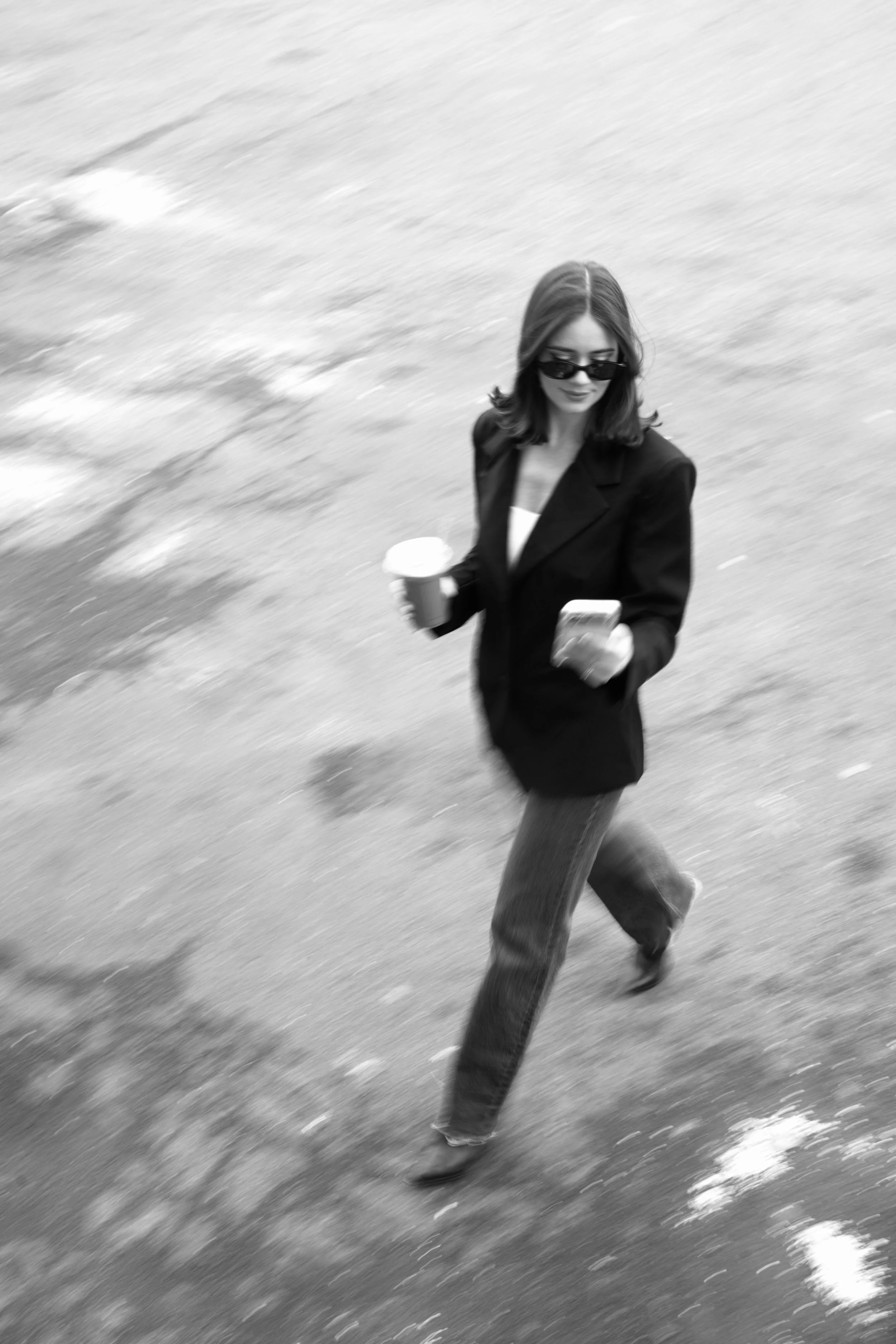 A woman walking on a sidewalk, holding a coffee cup in one hand and a phone in the other, wearing sunglasses, a blazer, and jeans.