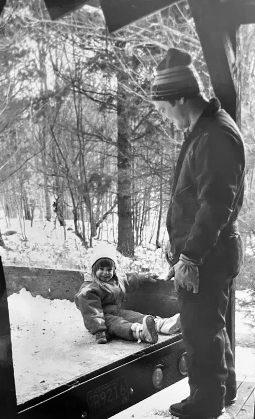 A young man and a little girl on a snow-covered outdoor deck during winter. The girl is sitting on the deck, smiling, while the young man stands beside her, holding her hand and looking down at her.