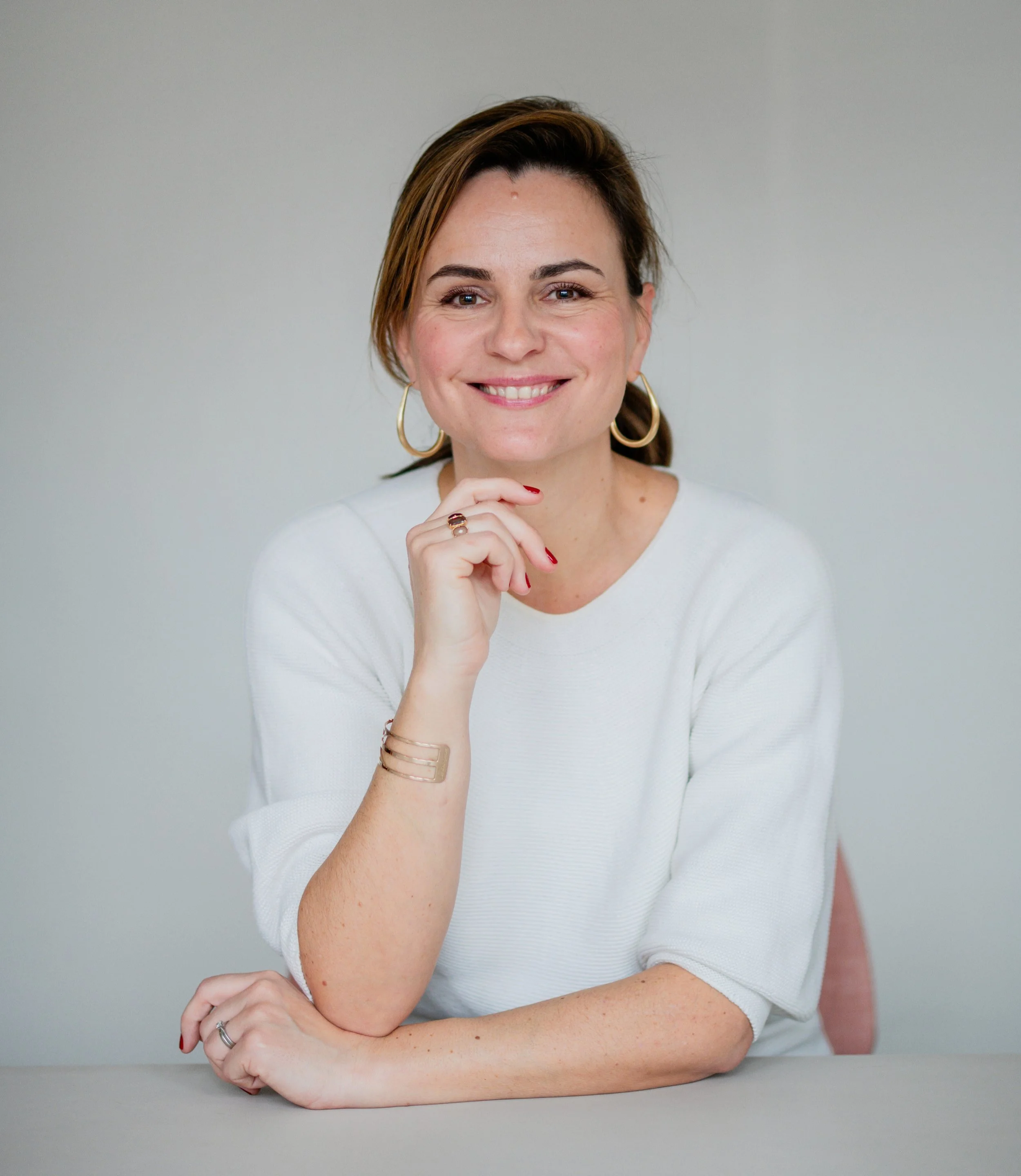 A woman with brown hair styled back, wearing hoop earrings, a white top, and a ring, sitting with her hand resting on her chin, smiling against a plain light gray background.