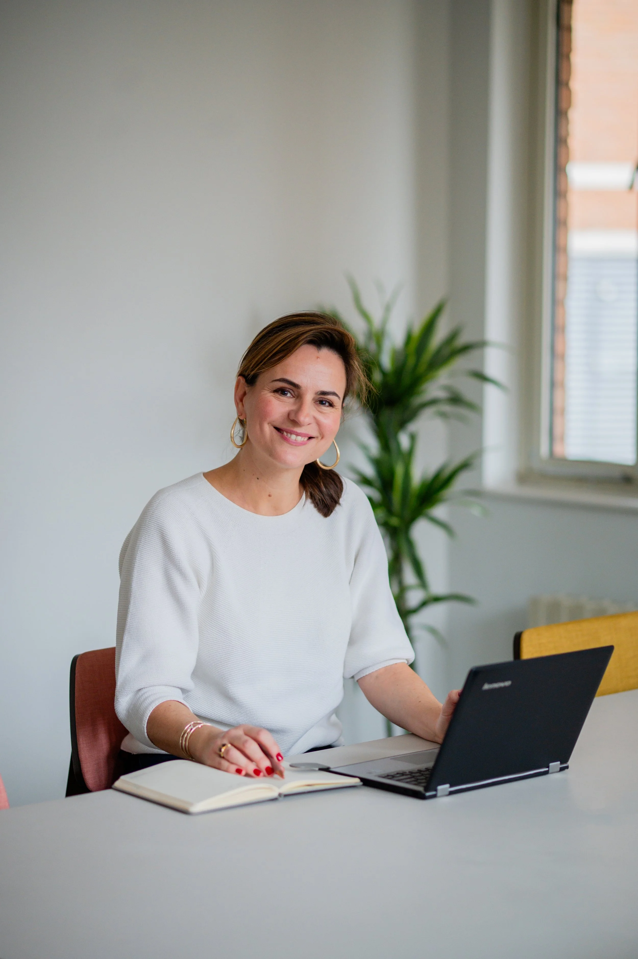 Smiling woman sitting at a desk with a laptop, notebook, and pen, in a bright room with a plant and window.